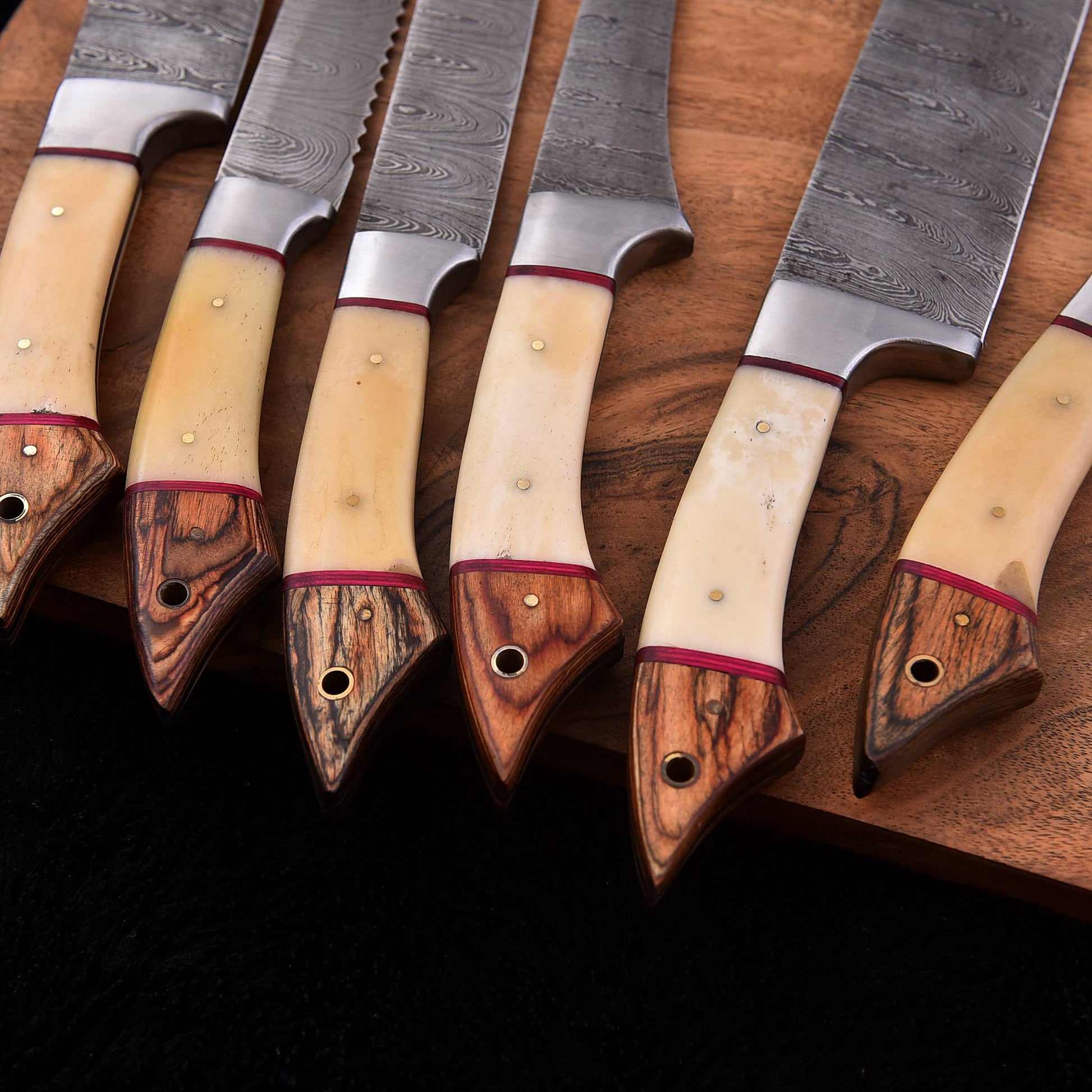 A set of kitchen knives with patterned steel blades, cream-colored bone handles, and dark wood ends, arranged diagonally on a wooden cutting board.