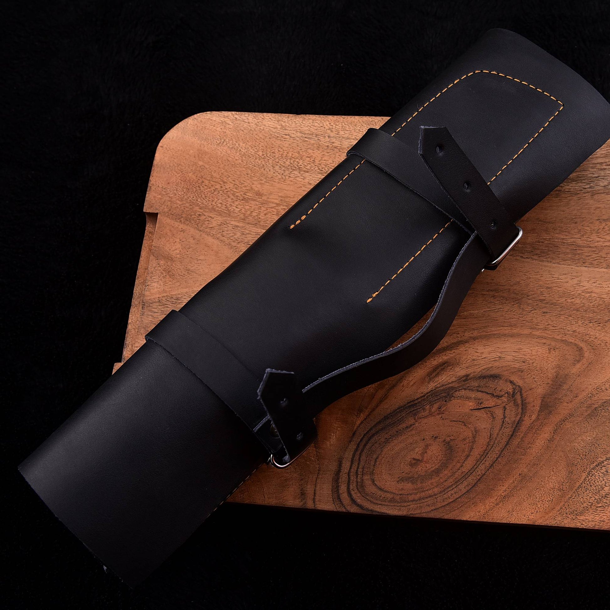 A black leather cylindrical case with a strap and buckle, laid diagonally on a wooden board against a black background.