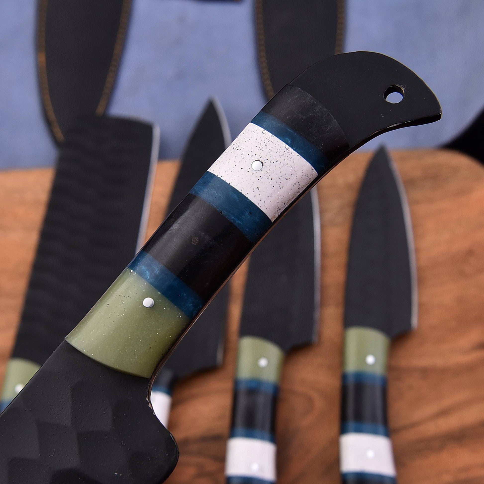 A close-up of a kitchen knife handle with a black ferrule, olive green section, blue stripe, and white speckled band, with other knives blurred in the background on a wooden surface.