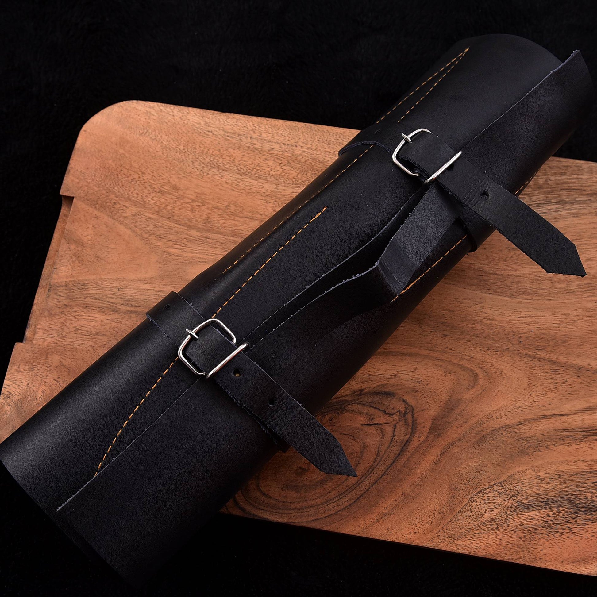 A black leather cylindrical case or roll-up pouch with two silver buckle straps, photographed diagonally on a wooden board against a black background.