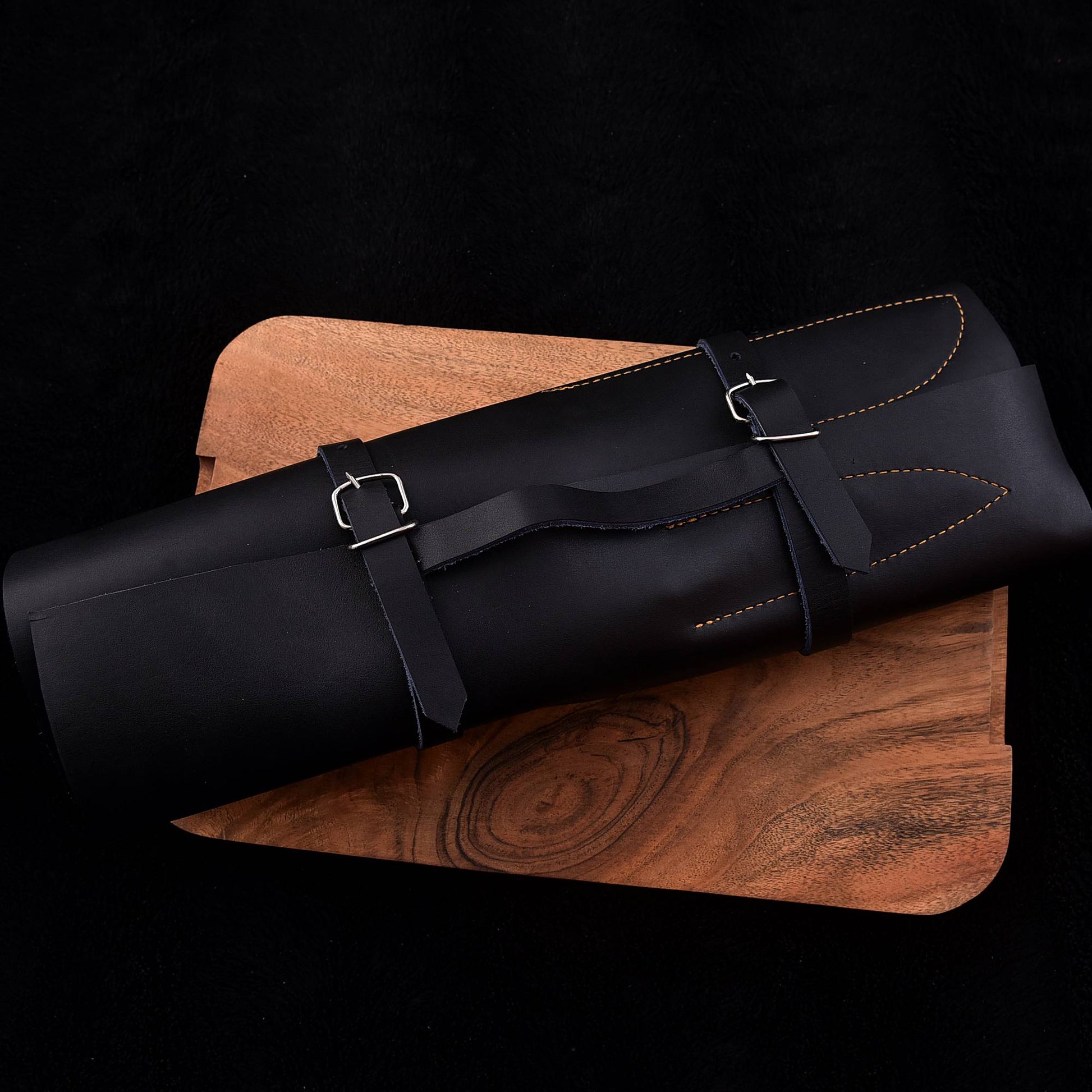 A black leather cylindrical pouch or case with two adjustable strap closures and silver buckles, resting diagonally on a brown wooden board against a black background.