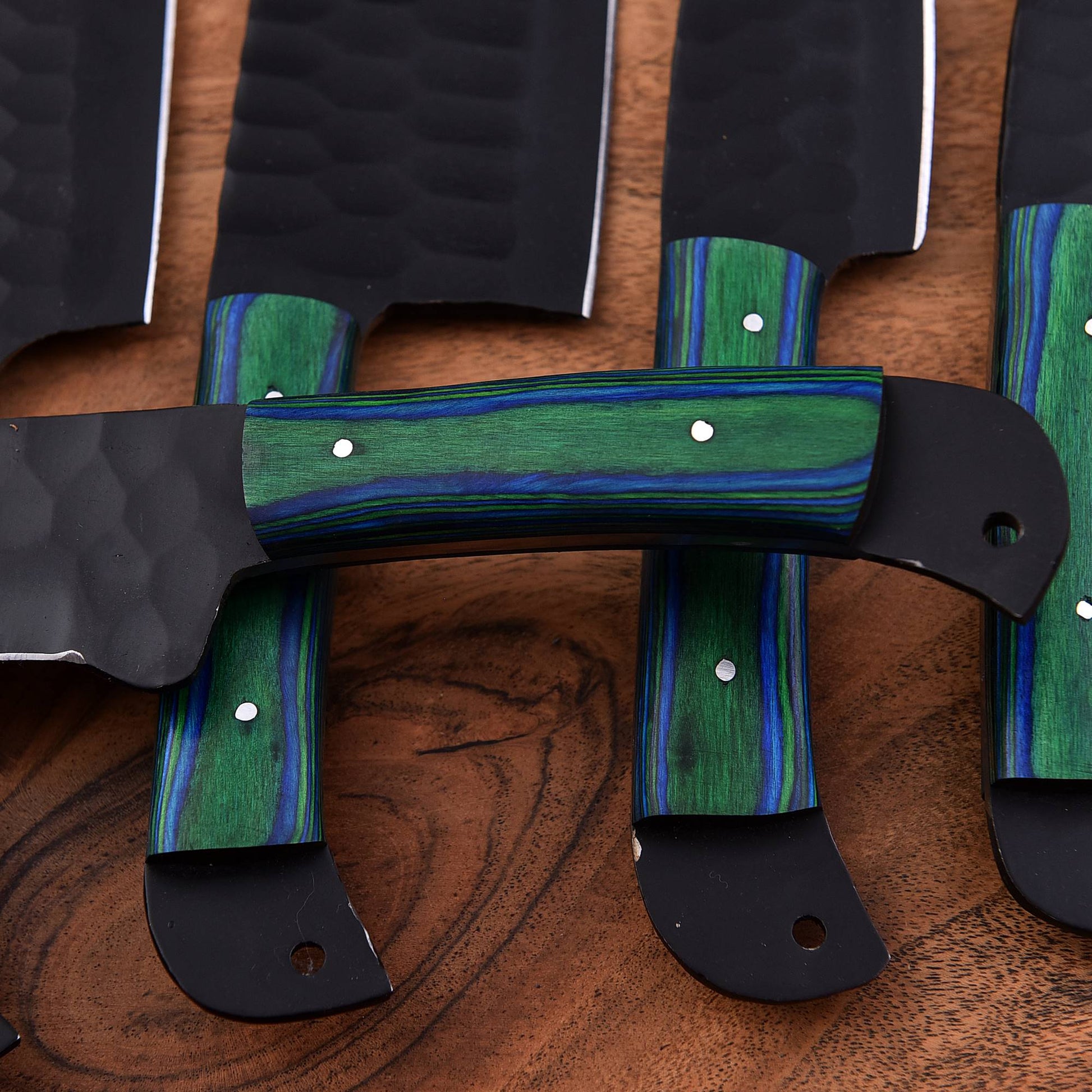 A close-up of a set of kitchen knives with textured black blades and handles featuring green and blue striped wood inlays, displayed on a wooden surface.