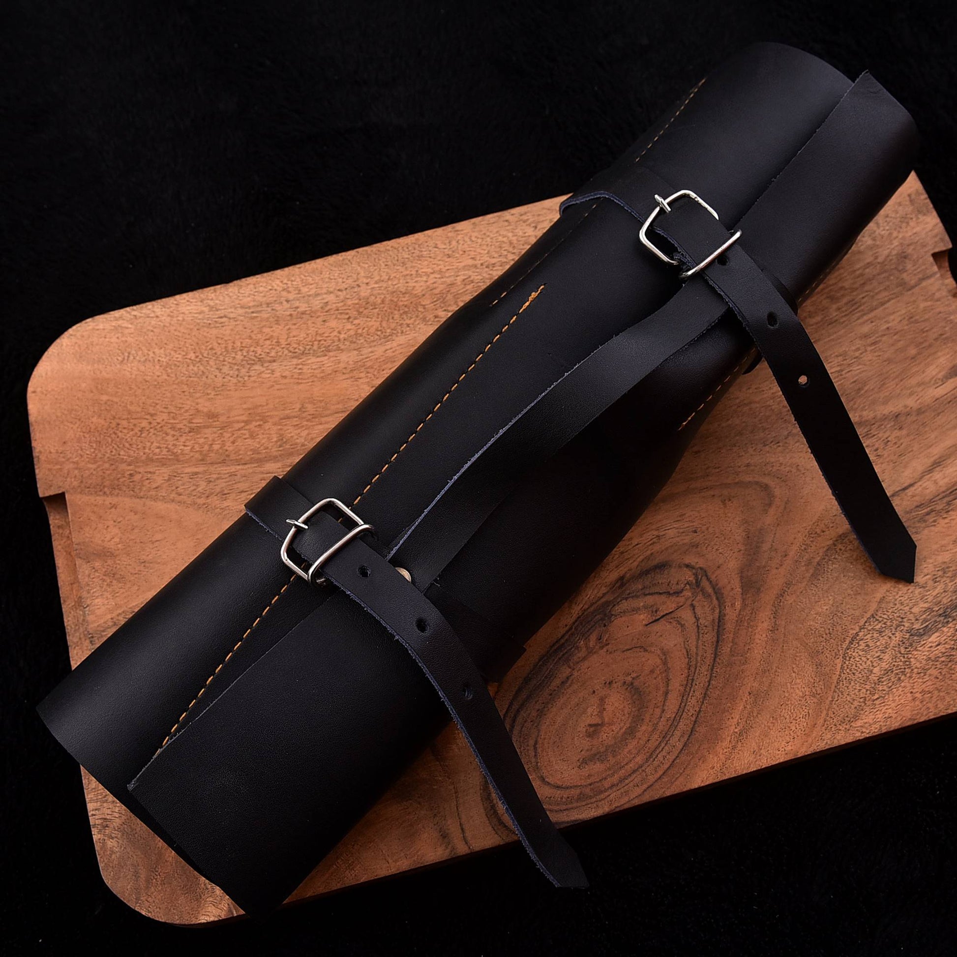 A black leather cylindrical roll-up strap or organizer with two silver buckle straps, laid diagonally on a wooden board against a black background.