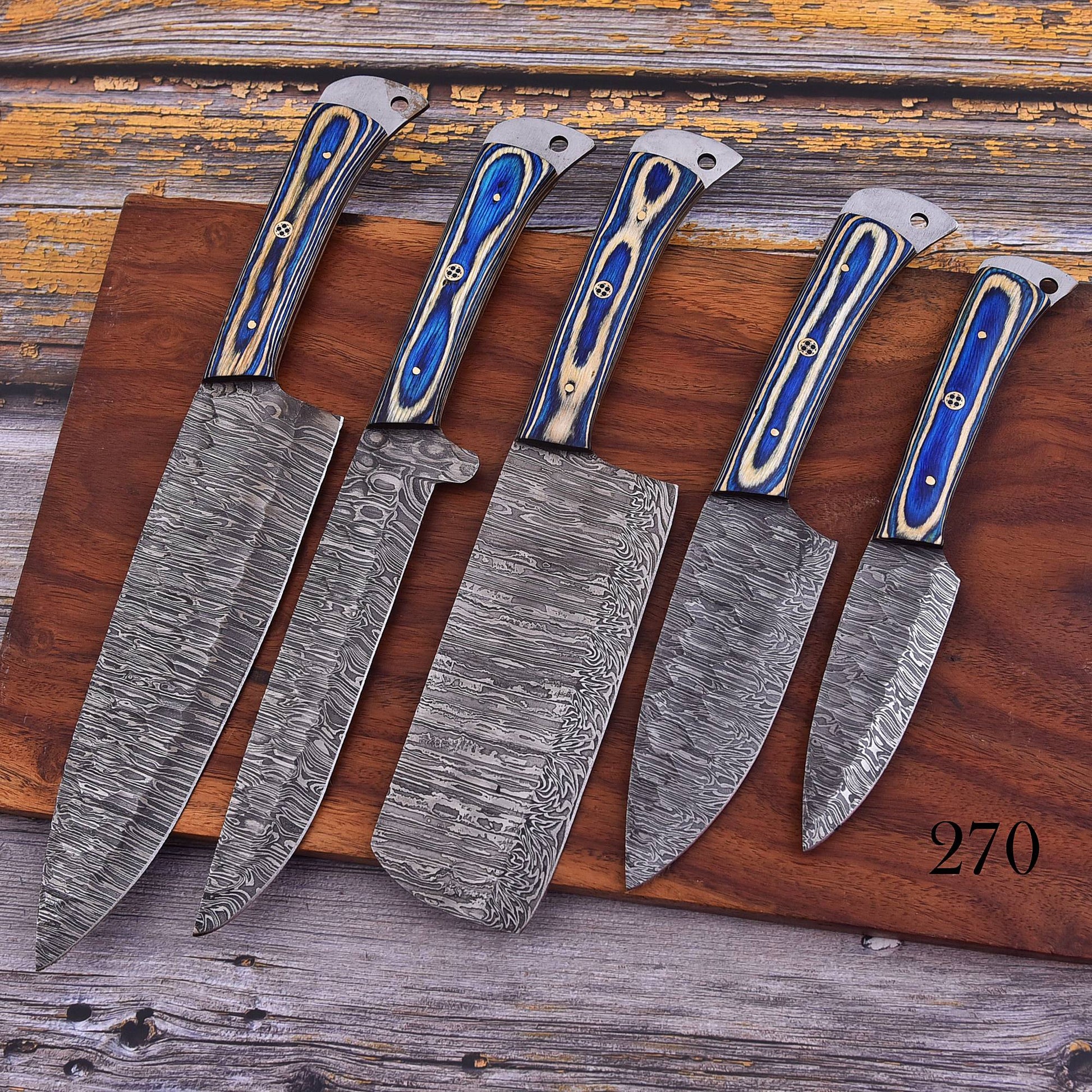Five Damascus steel kitchen knives of varying sizes arranged on a wooden cutting board over a rustic wooden surface. The knives feature patterned steel blades and handles with blue and beige inlay.