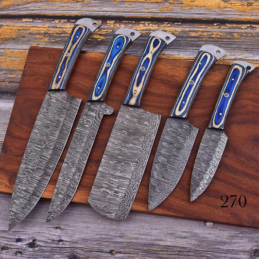 Five Damascus steel kitchen knives of varying sizes arranged on a wooden cutting board over a rustic wooden surface. The knives feature patterned steel blades and handles with blue and beige inlay.