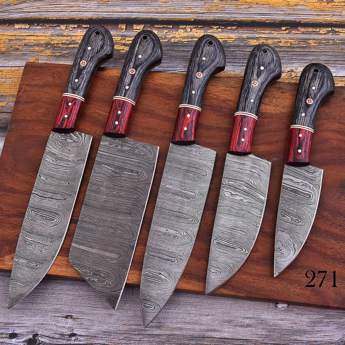 Five Damascus steel kitchen knives with dark wood handles and red ferrules, arranged diagonally on a wooden cutting board over a rustic wooden surface.