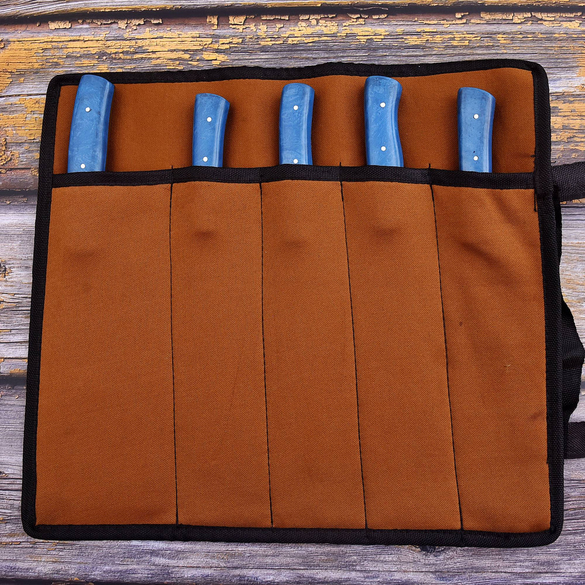 A brown fabric knife roll with black trim holding five knives with blue handles, laid flat on a weathered wooden surface.