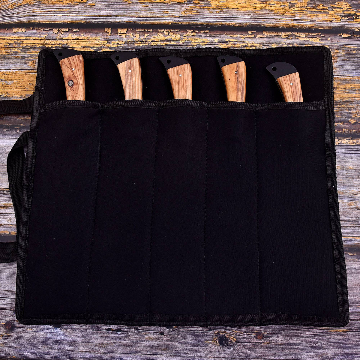 A black fabric knife roll holding five knives with light wood handles, laid flat on a rustic weathered wood surface.