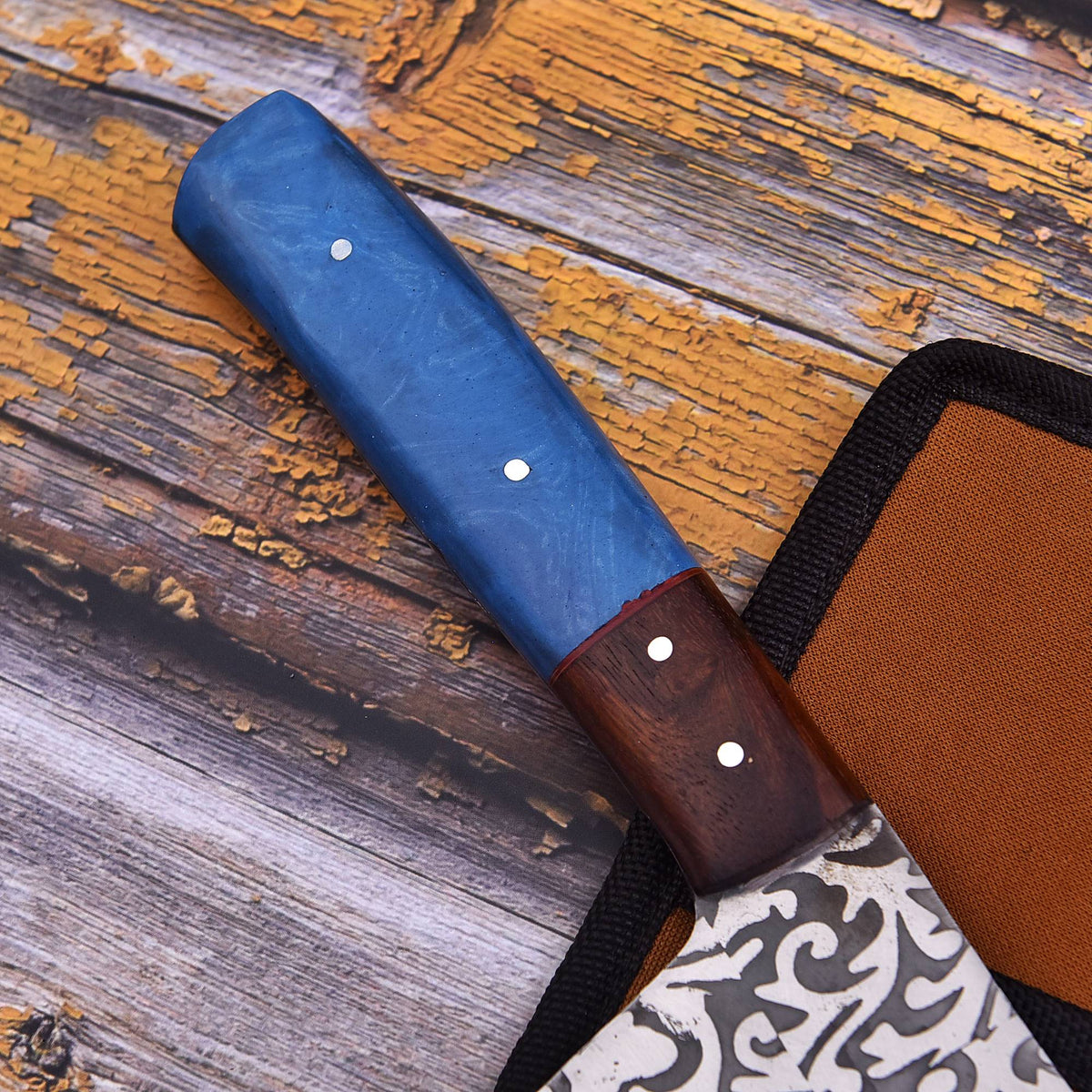 A close-up of a knife handle with a blue marbled section and a dark wood section, resting on a rustic wooden surface next to a brown sheath.