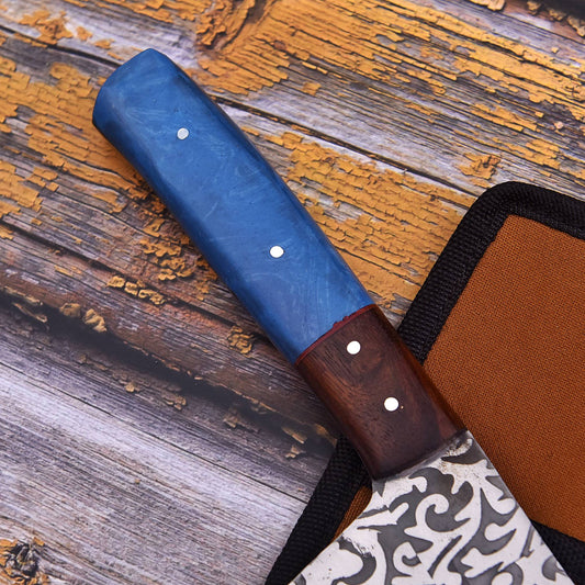 A close-up of a knife handle with a blue marbled section and a dark wood section, resting on a rustic wooden surface next to a brown sheath.