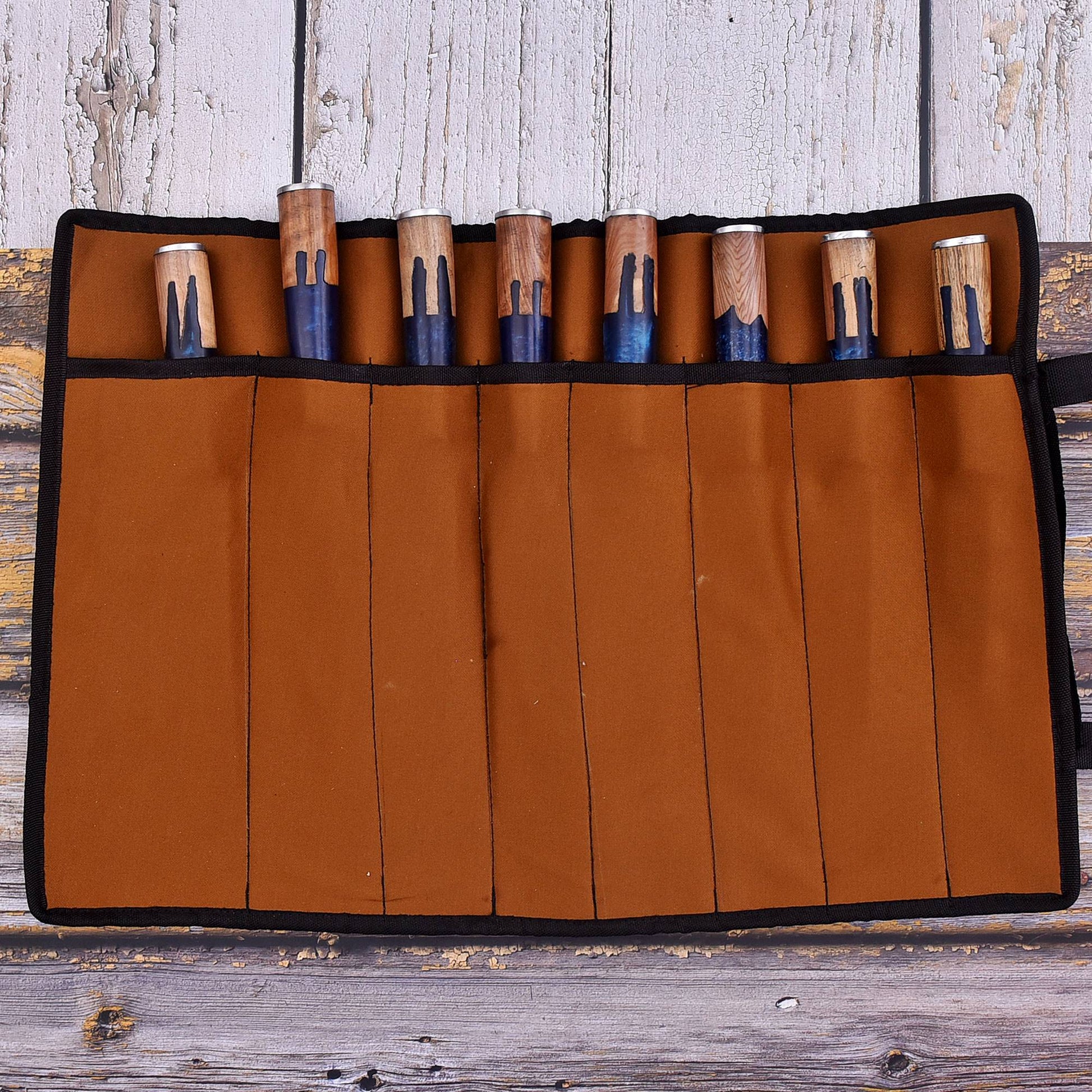 A brown canvas tool roll with black trim holding multiple wooden-handled tools with blue and white painted sections, laid flat on a rustic wooden surface.