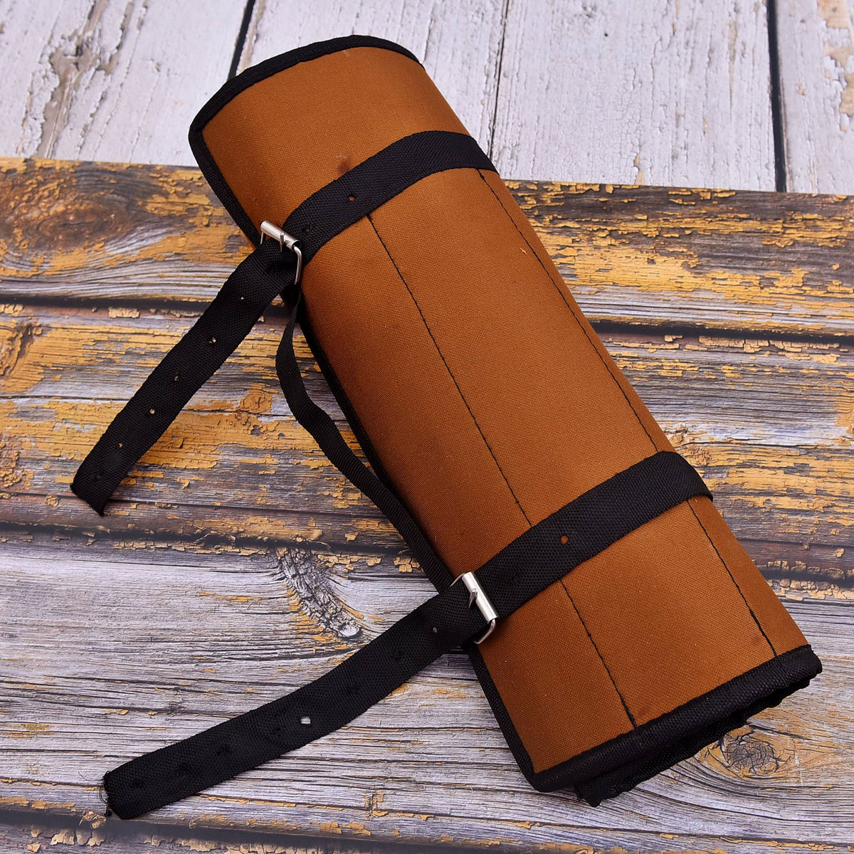A rolled brown leather or faux-leather strap holder with black adjustable straps and silver metal buckles, placed diagonally on a rustic weathered wood plank surface.