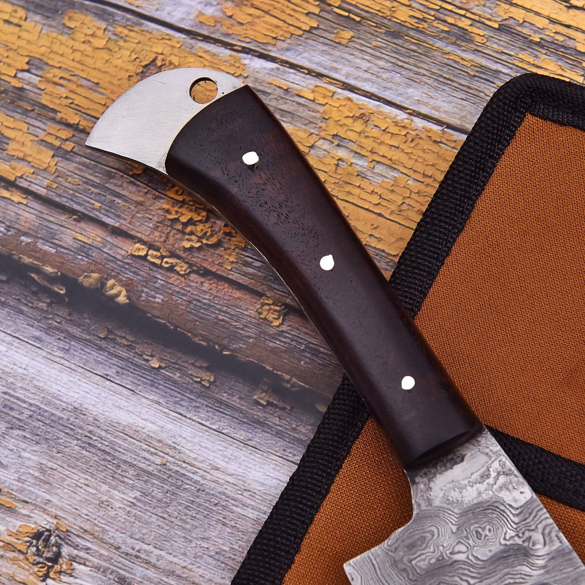 A close-up of a handcrafted knife with a dark brown wooden handle and a patterned steel blade, resting on a rustic weathered wood surface next to its orange and black sheath.
