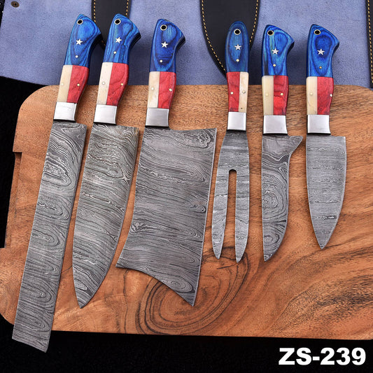 Seven Damascus steel kitchen knives with patriotic red, white, and blue handles are arranged on a wooden cutting board. The knives feature patterned steel blades and handles decorated with red, white, and blue sections and small white stars.