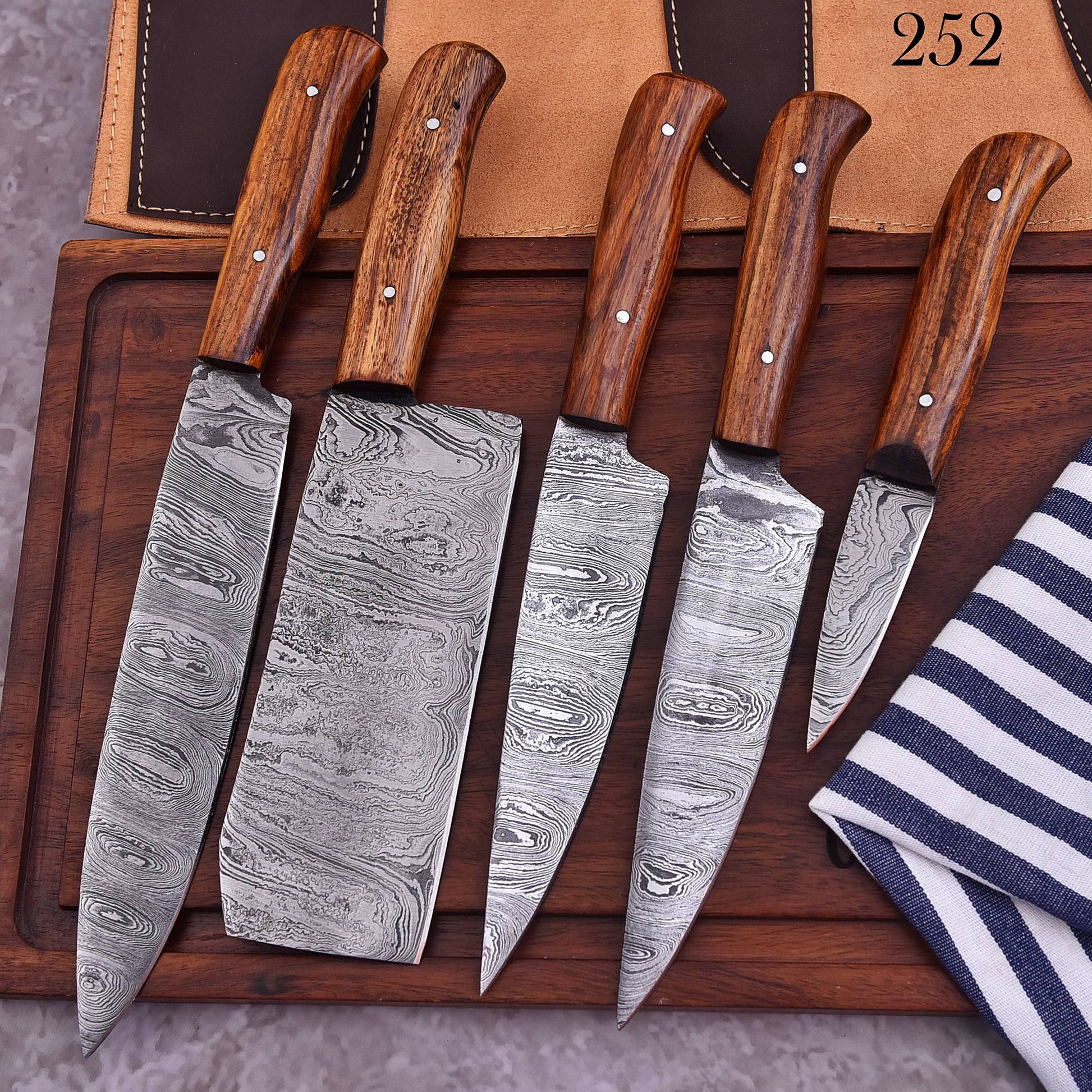 Five kitchen knives with patterned Damascus steel blades and brown wooden handles are arranged on a dark wooden board, with a tan leather sheath and a navy-and-white striped cloth in the background.
