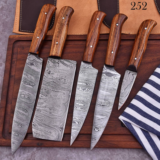 Five kitchen knives with patterned Damascus steel blades and brown wooden handles are arranged on a dark wooden board, with a tan leather sheath and a navy-and-white striped cloth in the background.