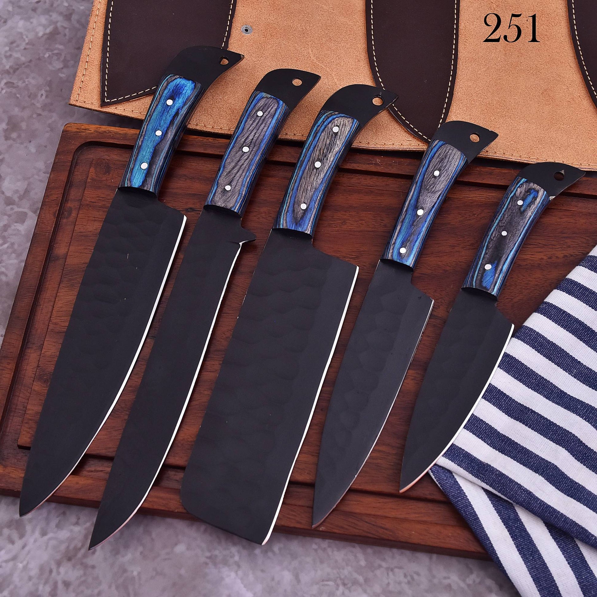Five kitchen knives with black blades and blue marbled wood handles arranged on a wooden board, with a tan knife roll and a striped cloth in the background.