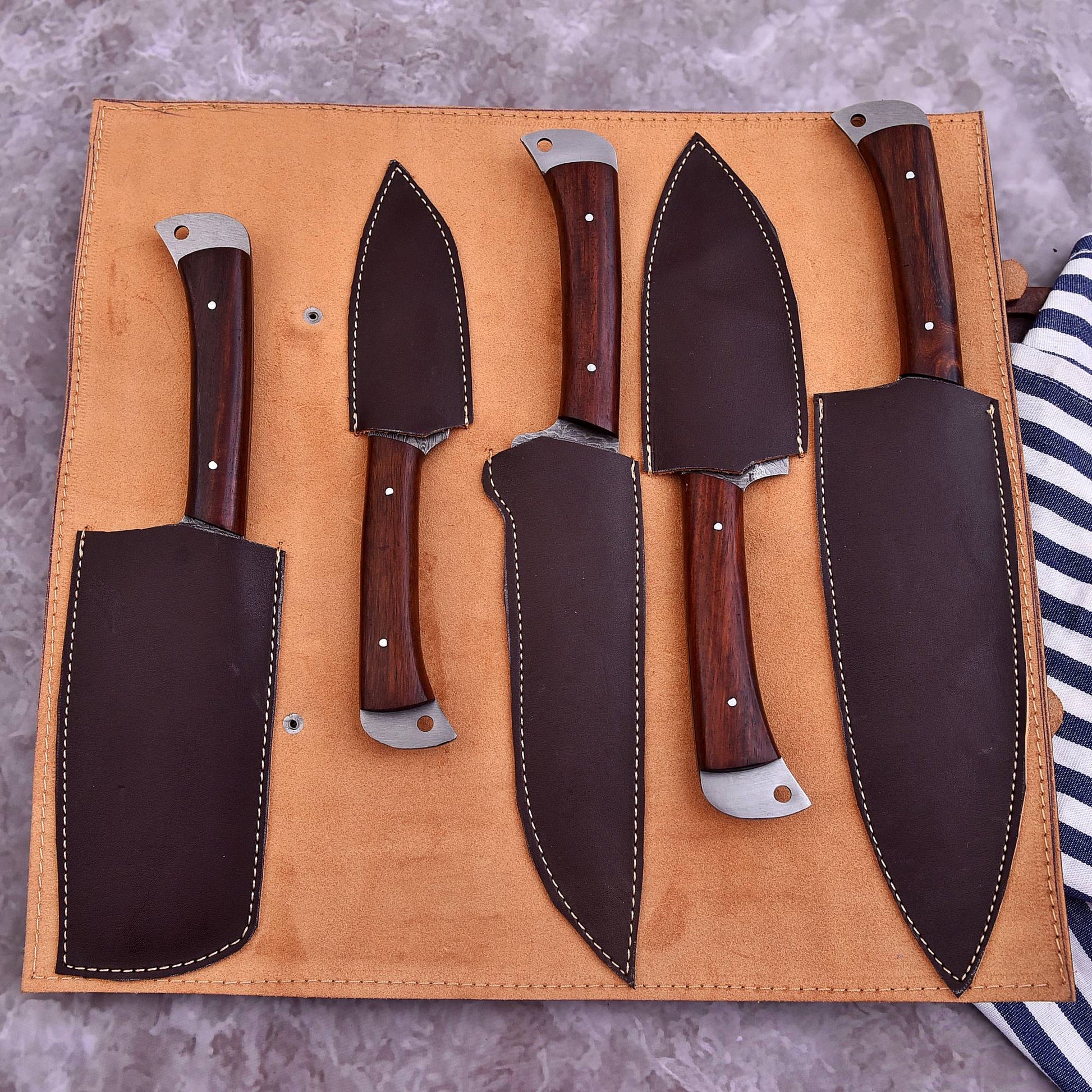 A set of five kitchen knives with dark wood handles and silver metal bolsters, arranged in a tan leather knife roll on a gray textured surface.