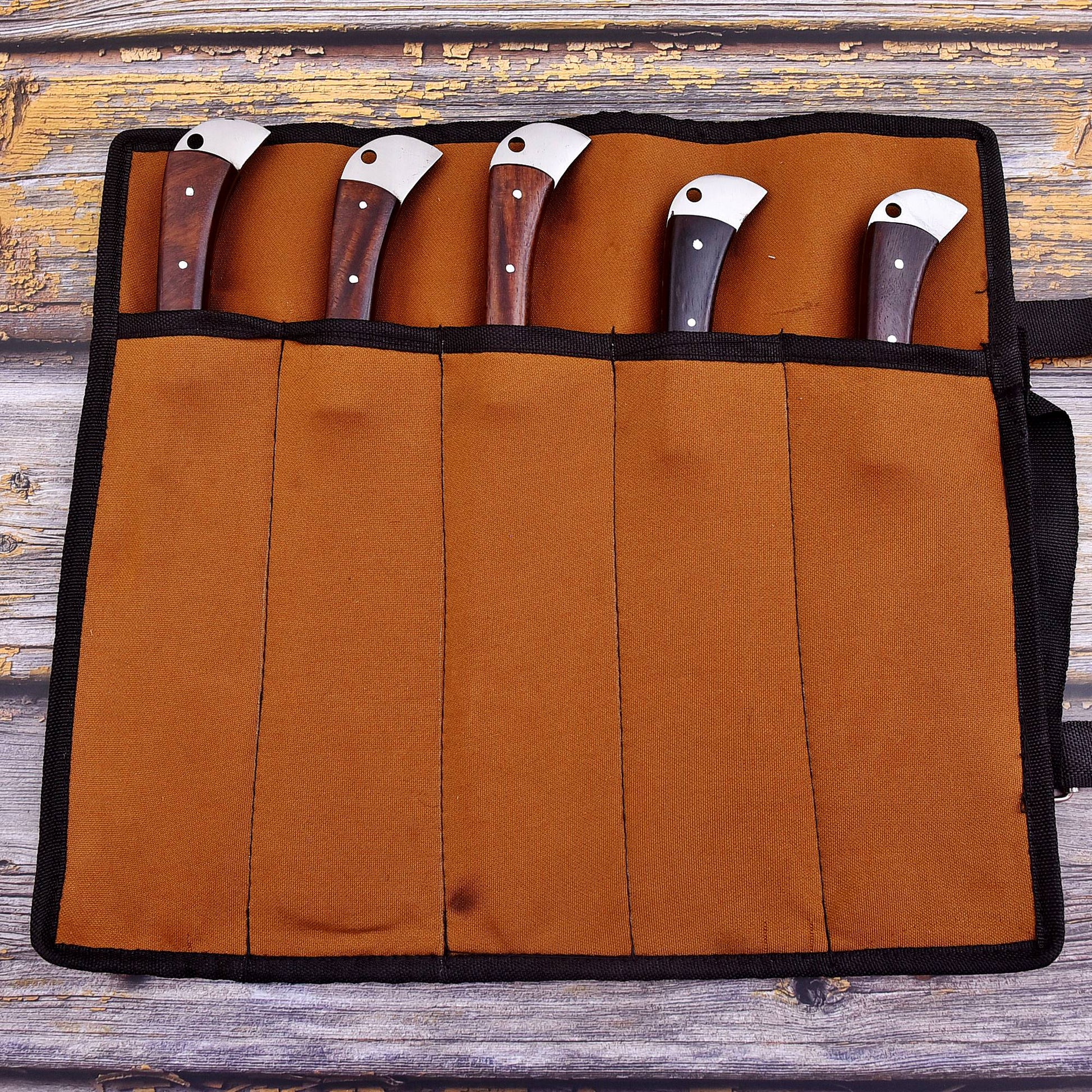 A brown suede knife roll with black trim holding five knives with dark wood handles and silver blades, laid flat on a rustic weathered wood background.