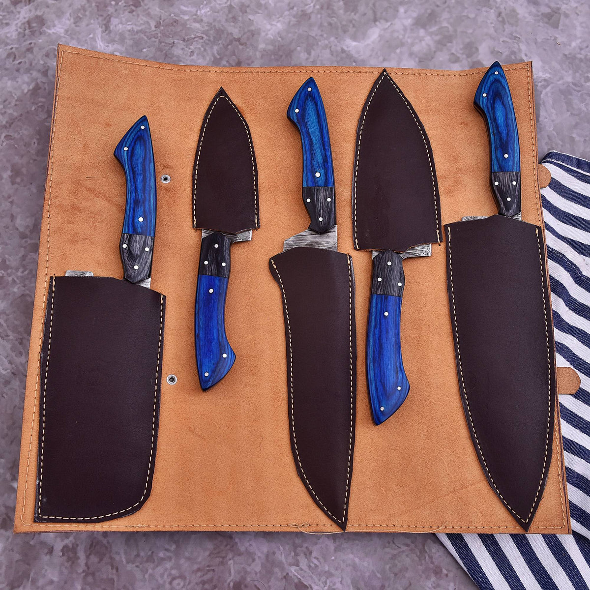 A set of five kitchen knives with vibrant blue wood handles and dark steel blades, presented in a tan leather roll-up organizer on a gray marbled surface.