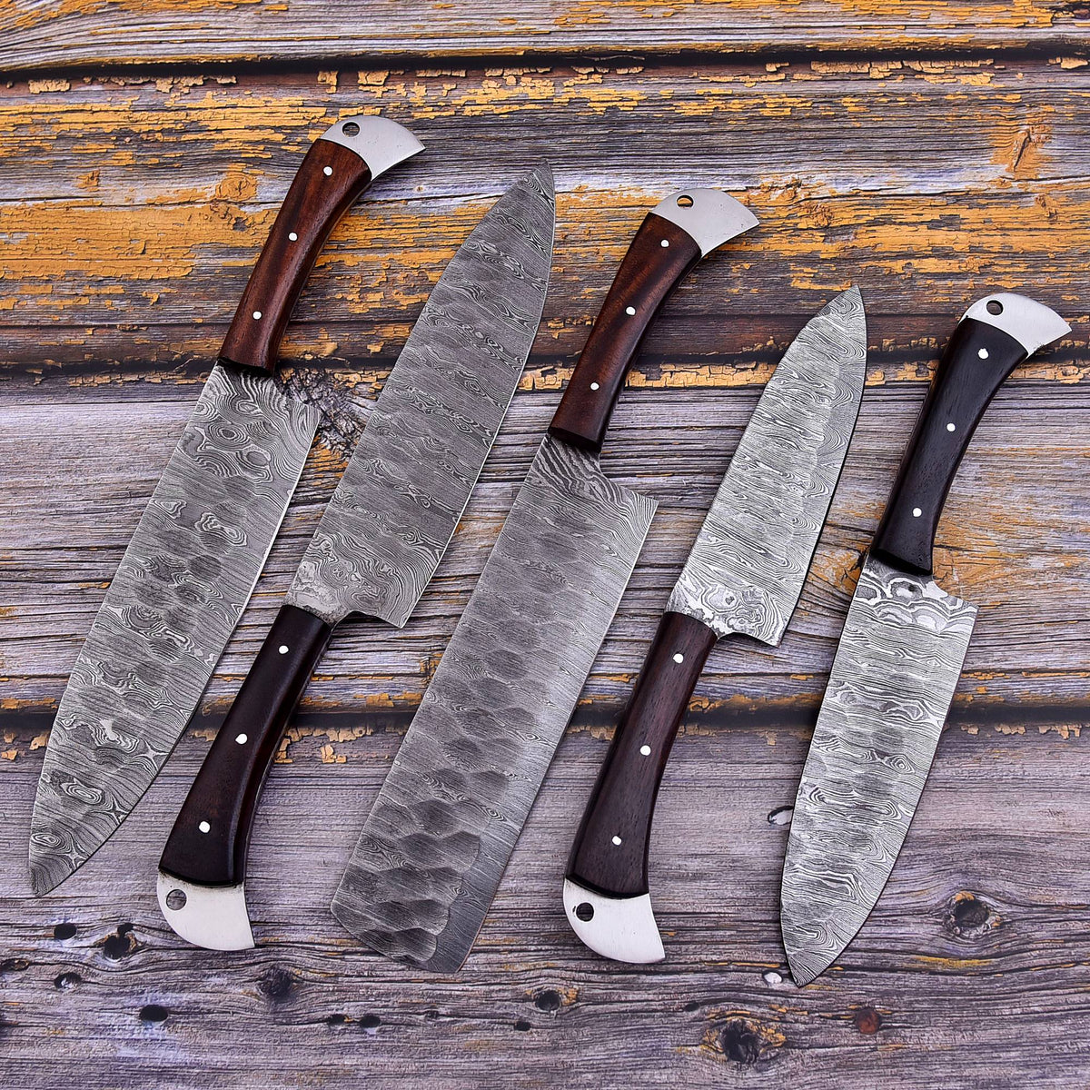 Four kitchen knives with patterned steel blades and dark wooden handles arranged diagonally on a rustic weathered wood background.