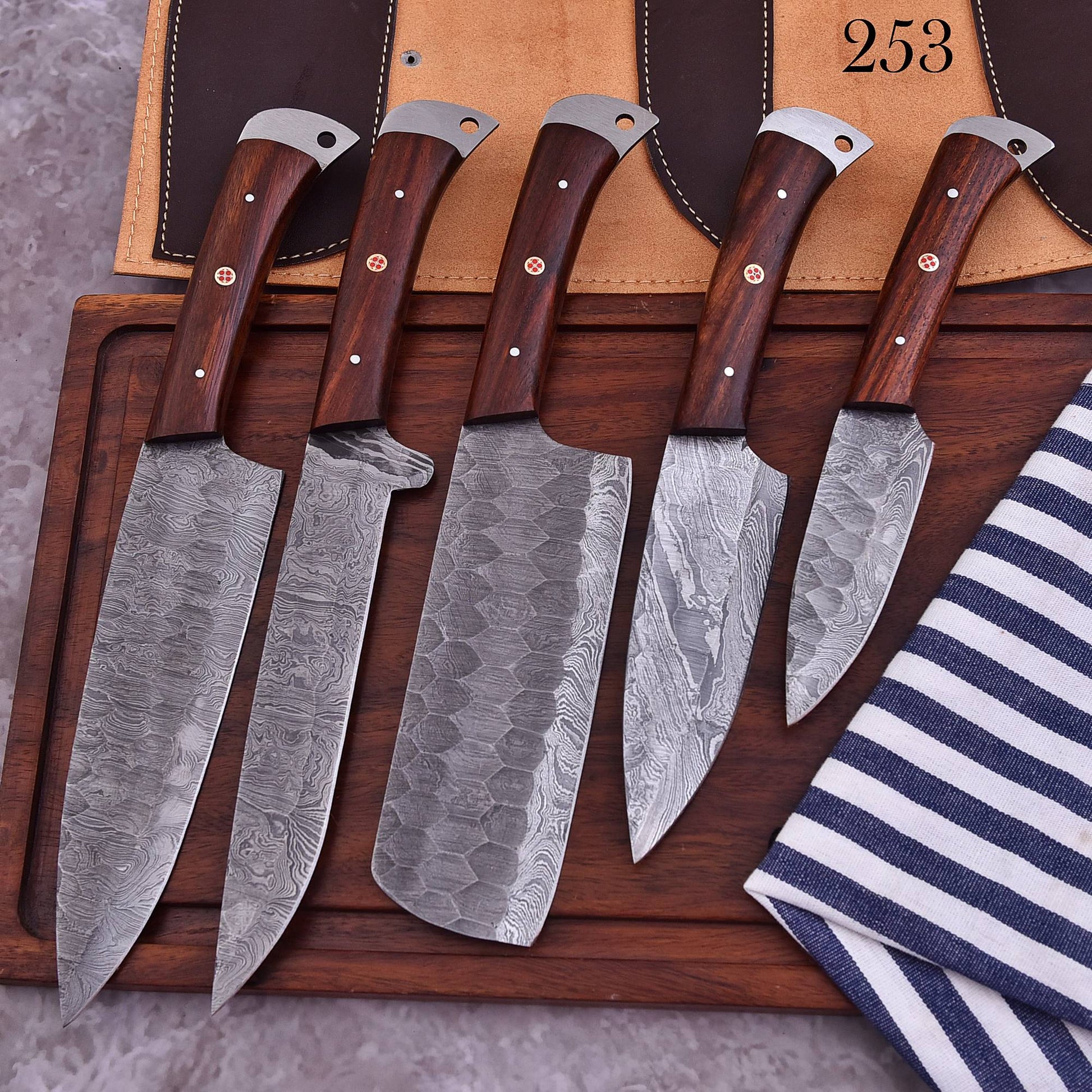 Five Damascus steel kitchen knives with dark wood handles are arranged on a wooden cutting board, with a tan knife sheath and a navy-and-white striped cloth visible in the background.