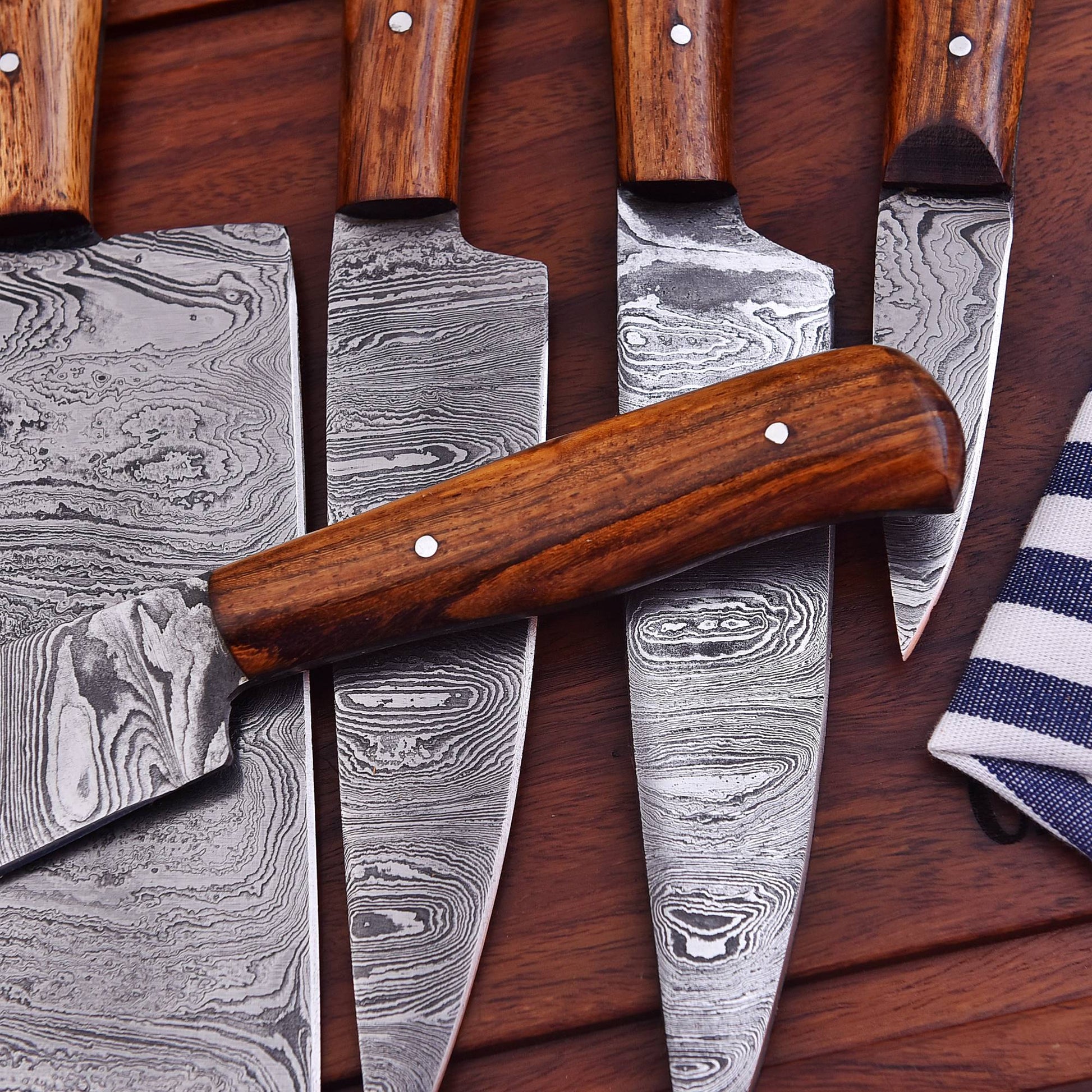 A set of kitchen knives with patterned Damascus steel blades and brown wooden handles, arranged on a dark wood surface with a striped cloth partially visible.
