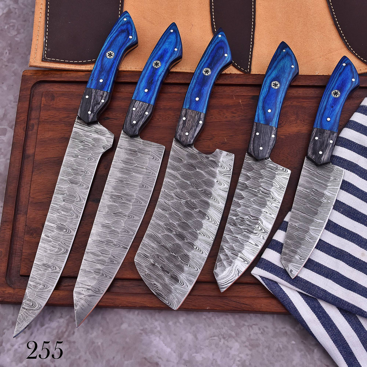 Five kitchen knives with patterned steel blades and blue wooden handles are arranged on a dark wooden board, with a tan leather sheath and a blue-and-white striped cloth nearby.