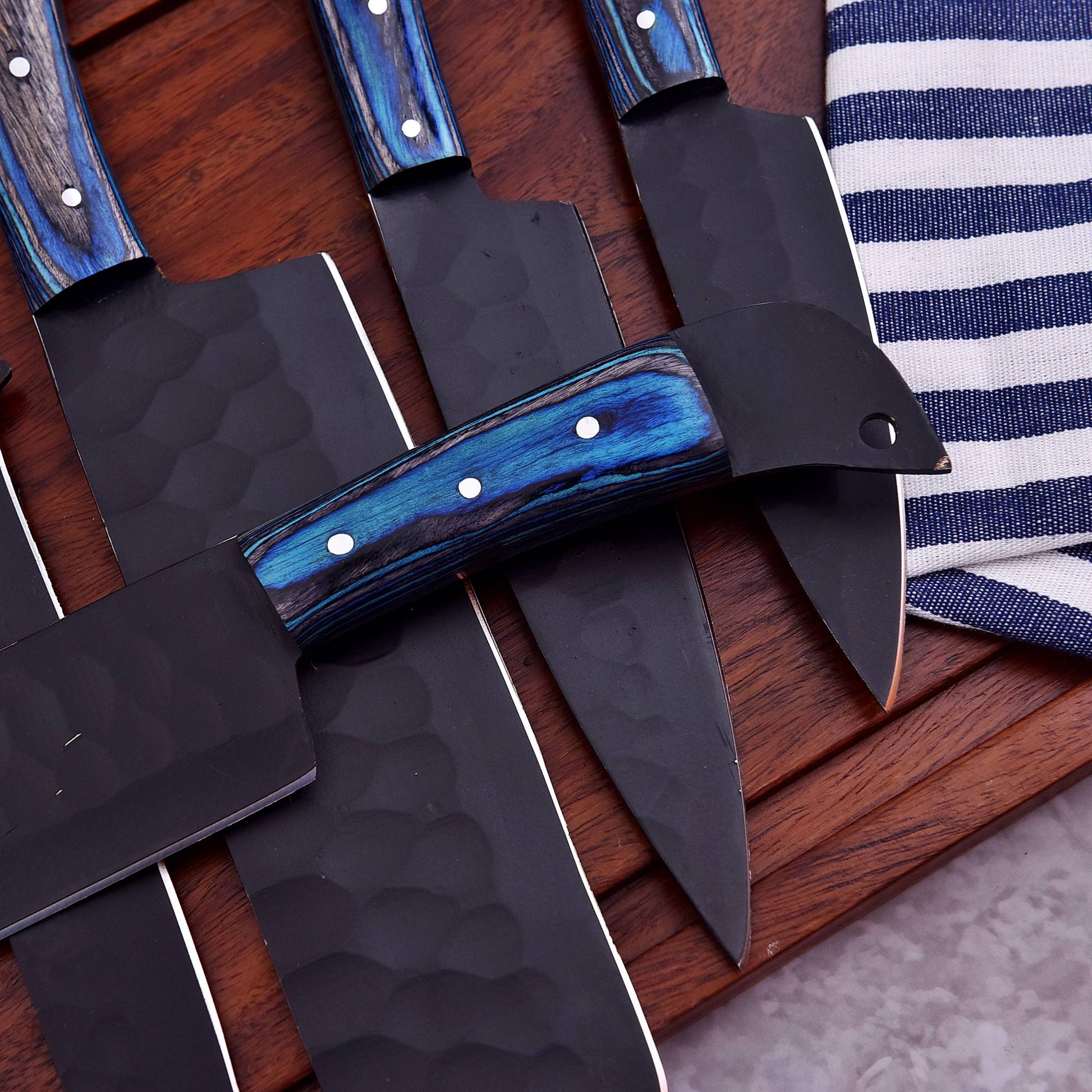 Four kitchen knives with black hammered blades and blue wood handles arranged on a wooden surface next to a striped cloth.