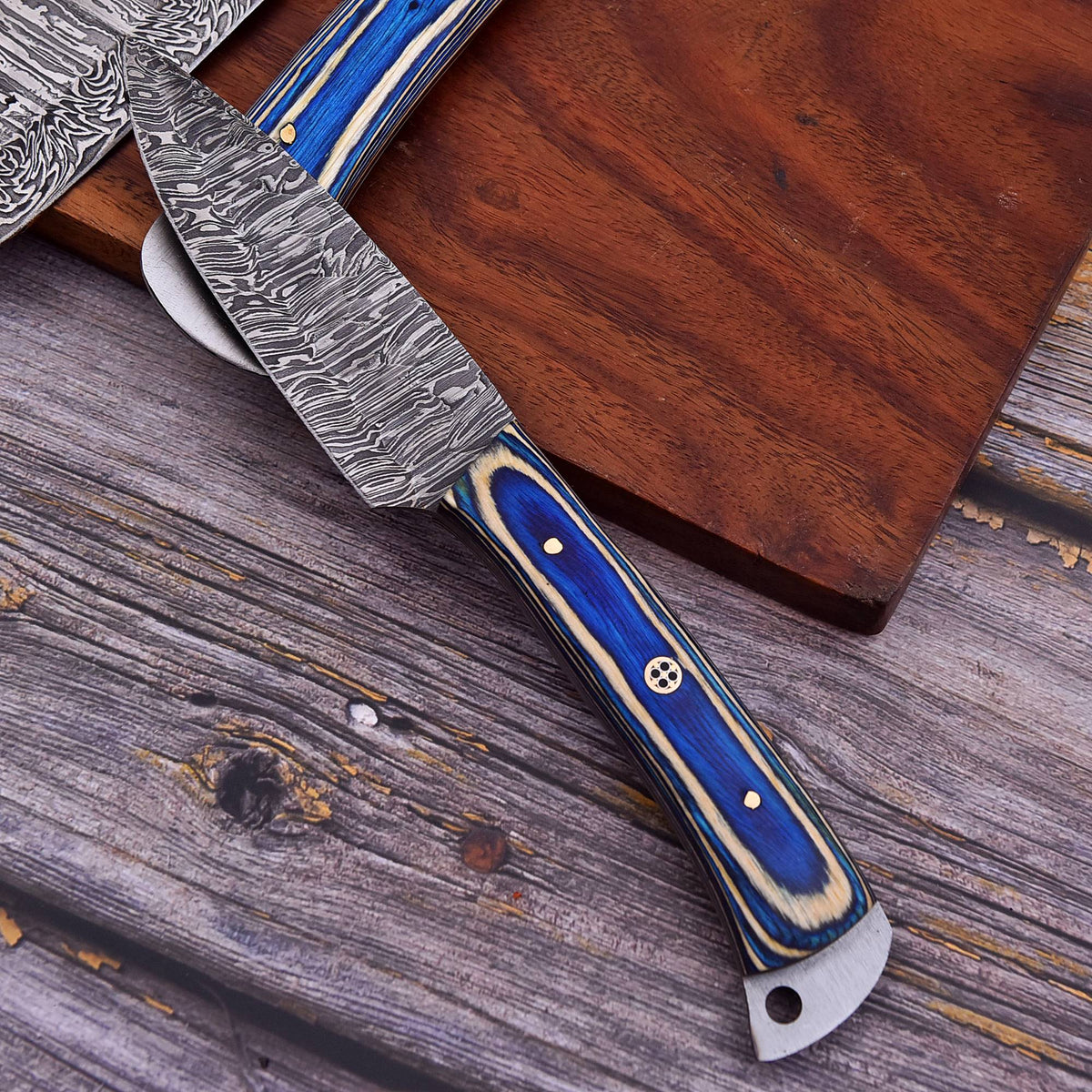 A close-up of a fixed-blade knife with a patterned steel blade and a blue-and-cream handle, resting on a wooden cutting board over a weathered wood surface.