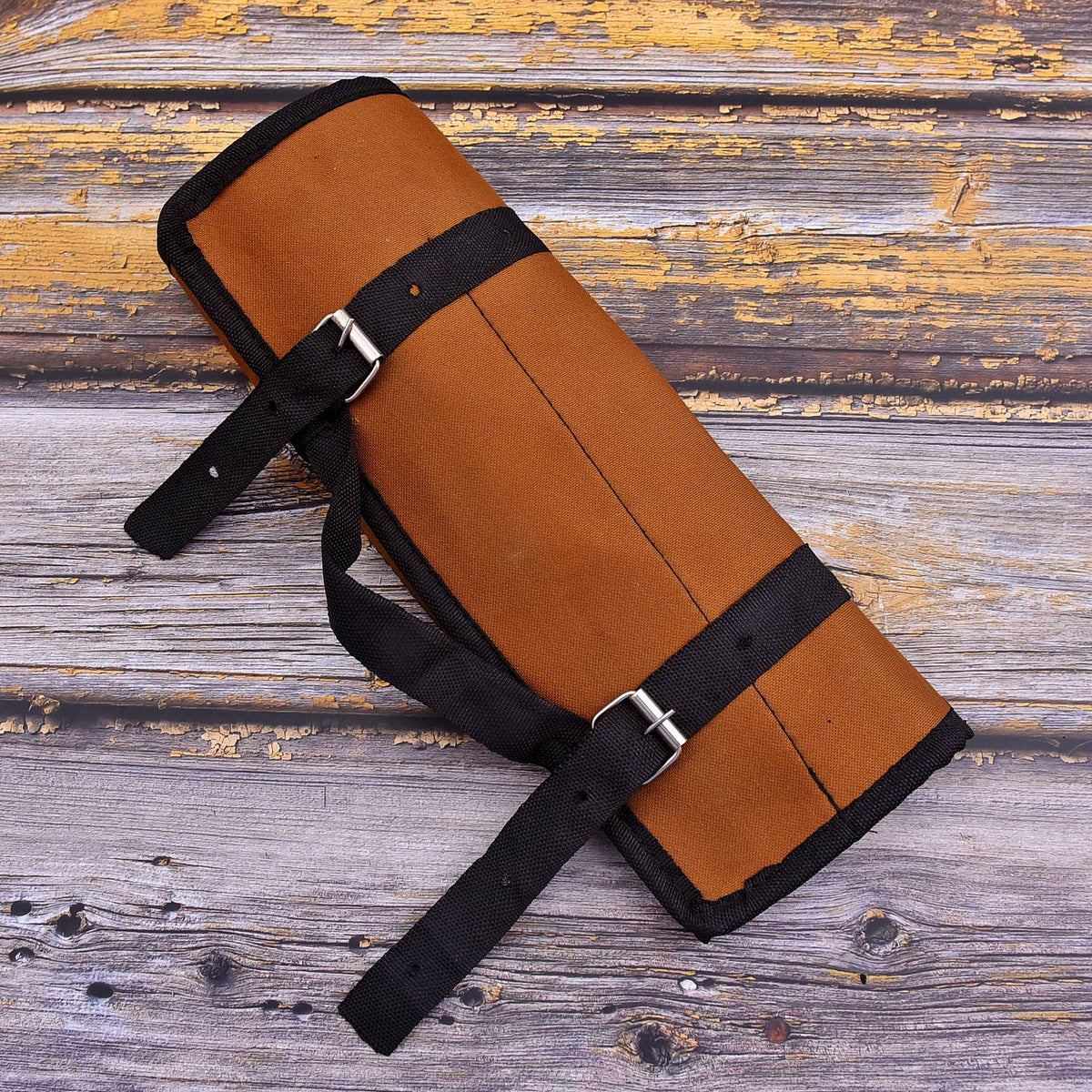 A rolled tan leather protective pad or guard with black straps and silver metal buckles, laid diagonally on a rustic weathered wood plank surface.
