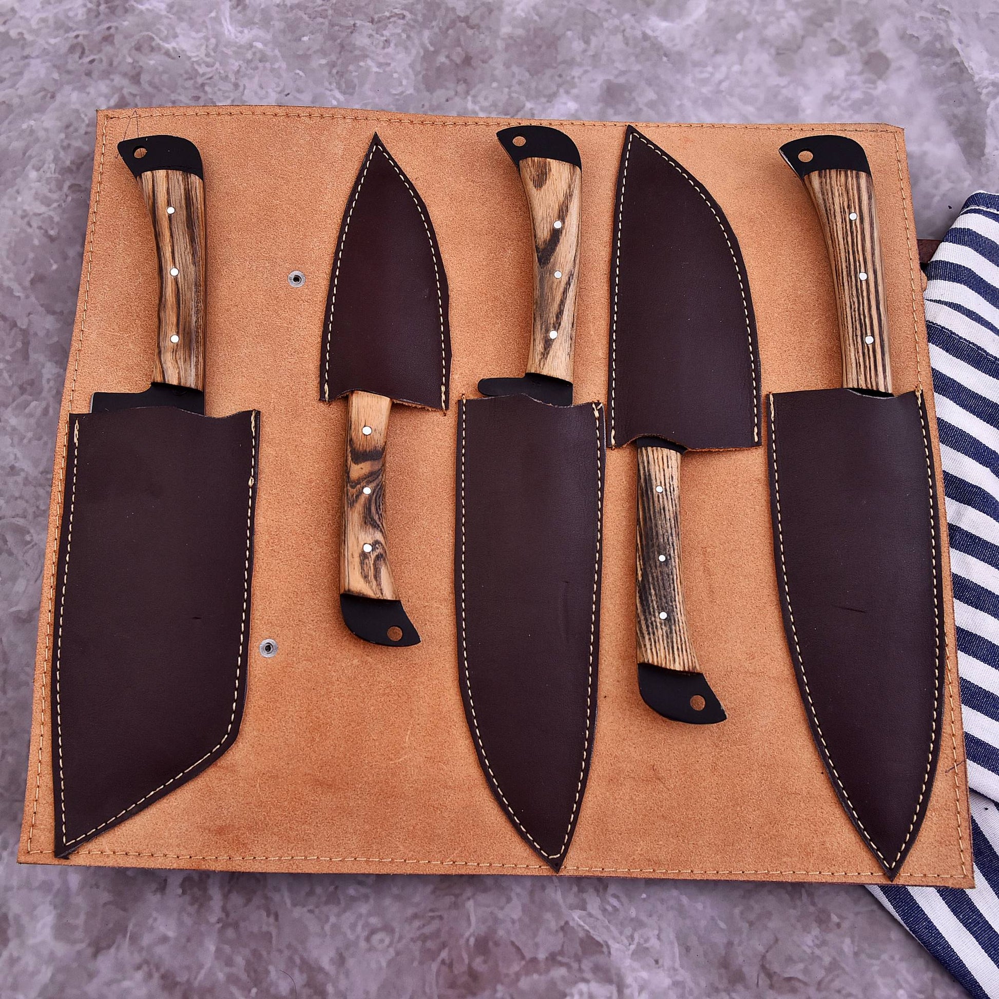 A set of four kitchen knives with natural wood handles and dark brown leather sheaths, arranged inside a tan leather knife roll on a gray stone surface.