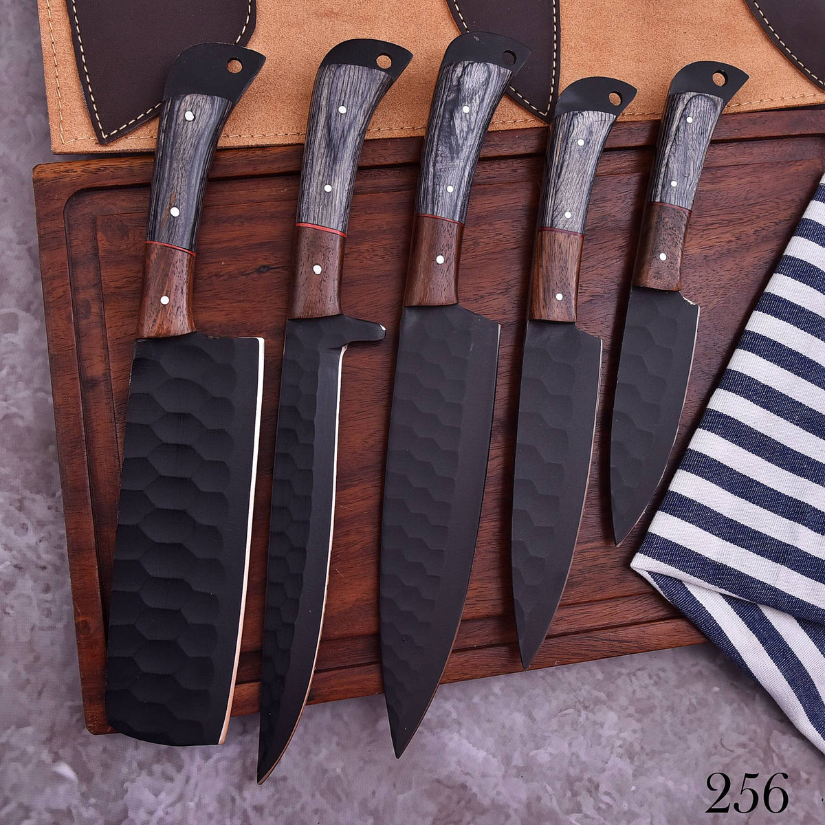Five kitchen knives with black hammered blades and wood-and-stone handles are arranged on a dark wooden board, with two matching black sheaths partly visible behind them. A striped cloth and gray textured surface surround the setup.