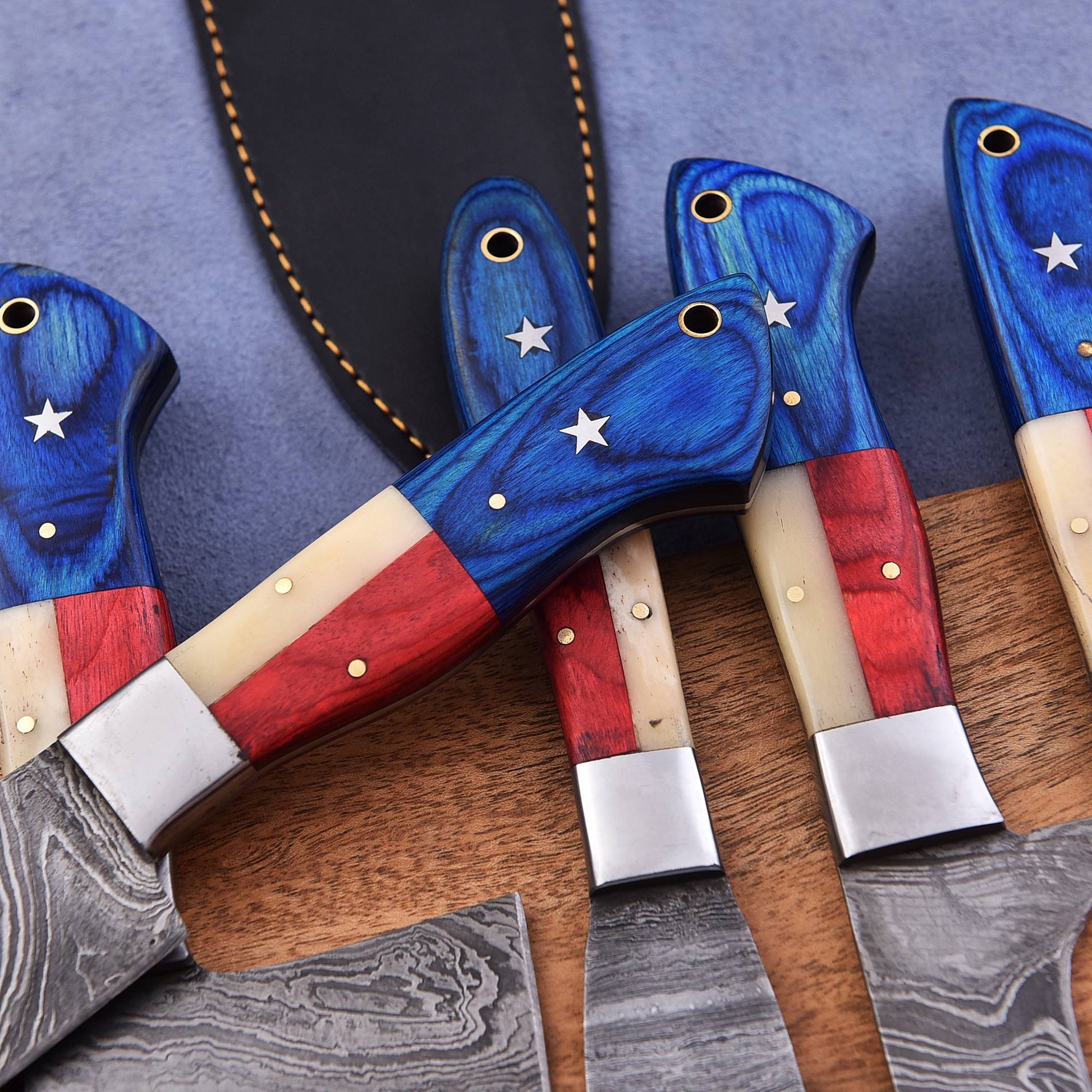 Several fixed-blade knives with decorative handles are arranged on a wooden surface. The handles feature a blue top section with white stars, red and cream stripes, and silver metal bolsters.