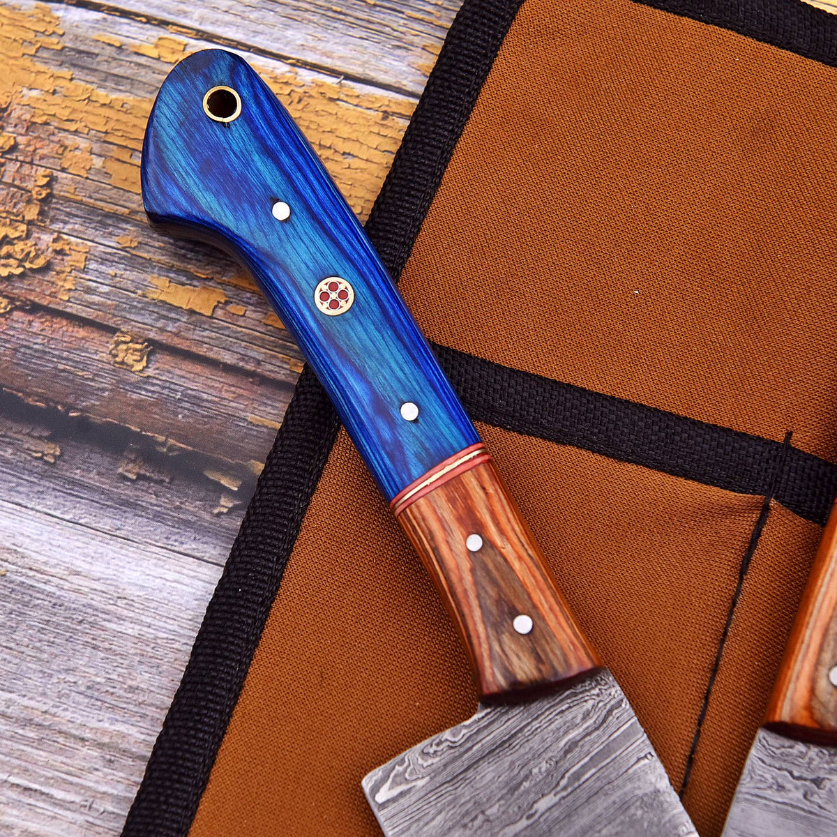 A close-up of a handcrafted knife with a vibrant blue wood handle and a Damascus steel blade, resting on a brown canvas knife roll over a rustic wooden surface.