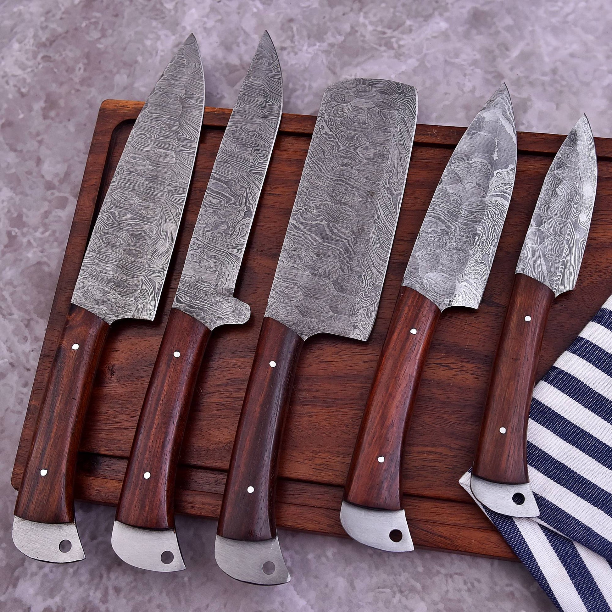 Five Damascus steel kitchen knives with dark brown wooden handles arranged on a wooden cutting board over a gray stone surface, with a striped cloth partially visible at the lower right.
