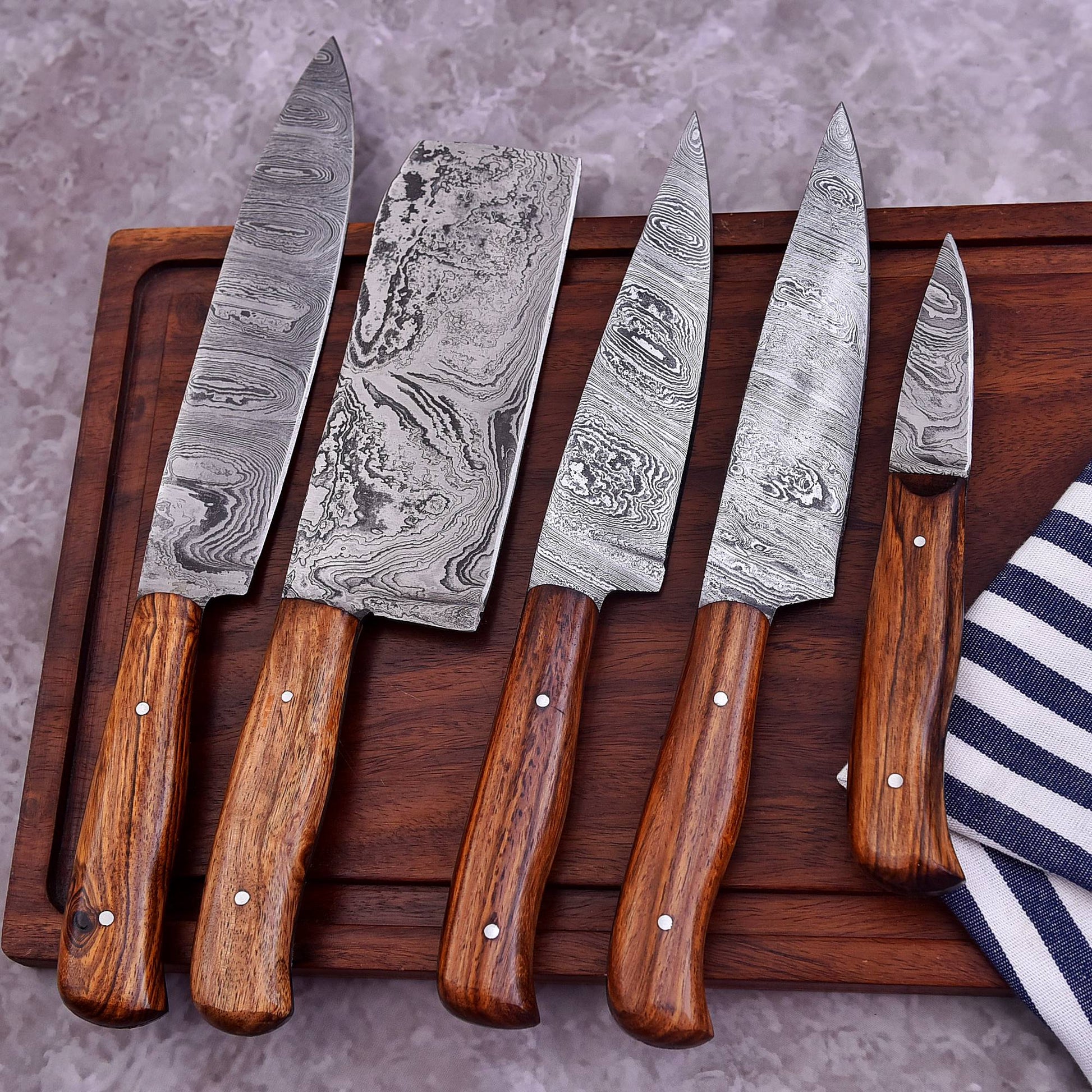 Five kitchen knives with patterned Damascus steel blades and brown wooden handles arranged on a dark wooden cutting board, with a striped cloth partially visible on the right.