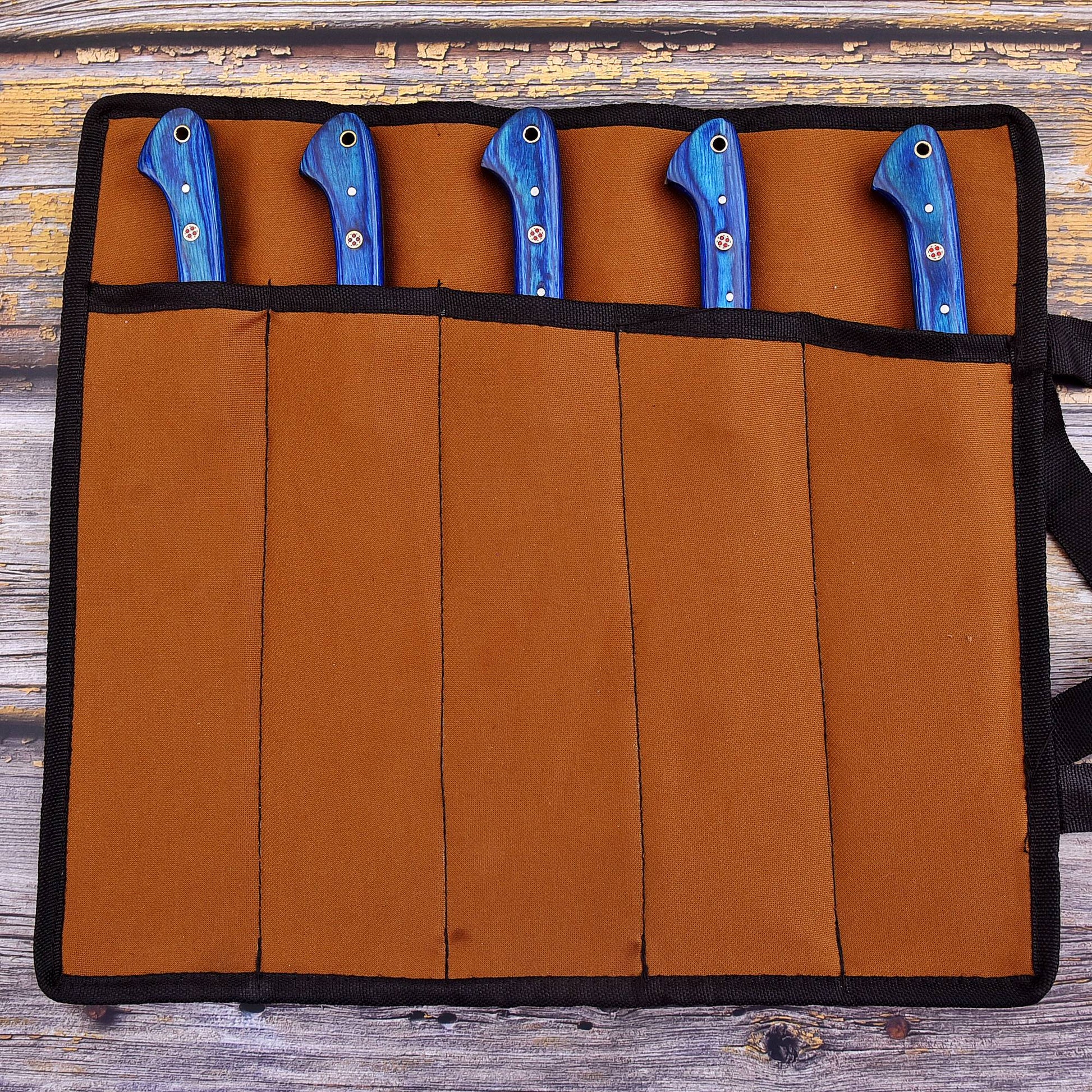 A brown fabric knife roll with black trim holding five knives with blue handles, laid flat on a rustic wooden surface.