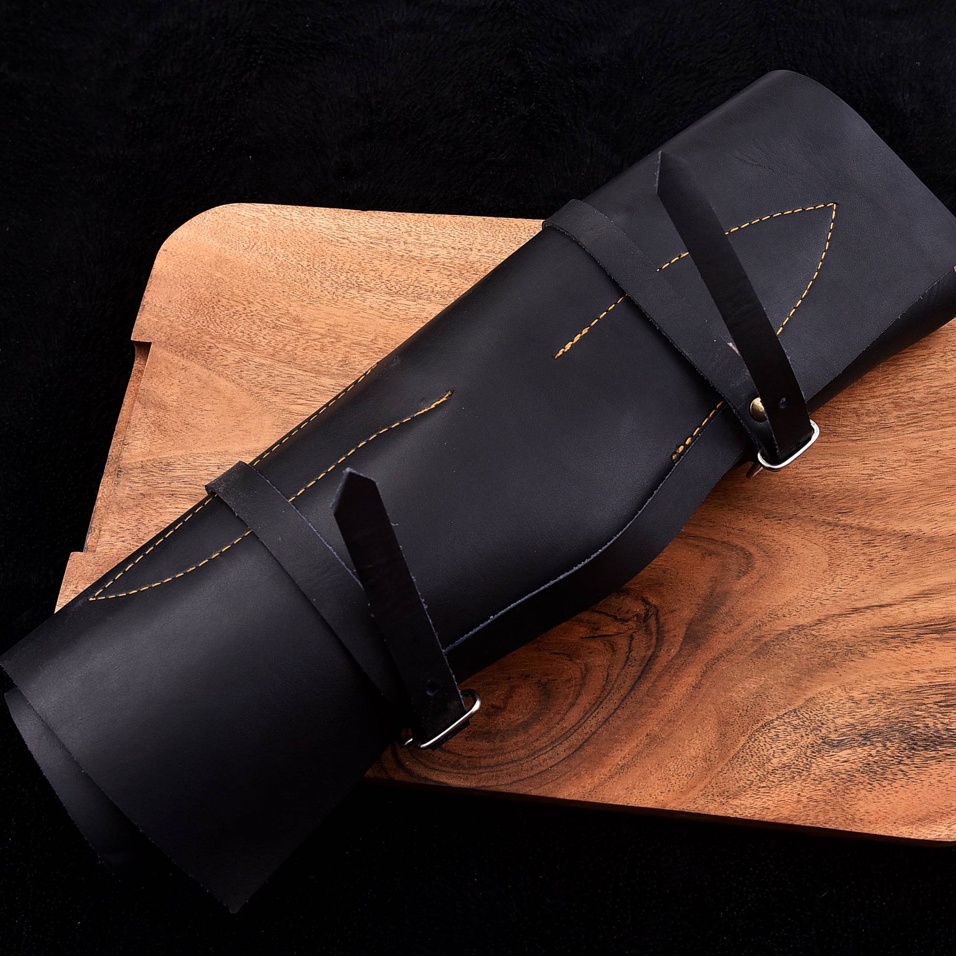 A black leather knife roll with adjustable straps and contrast stitching, resting diagonally on a wooden cutting board against a black background.