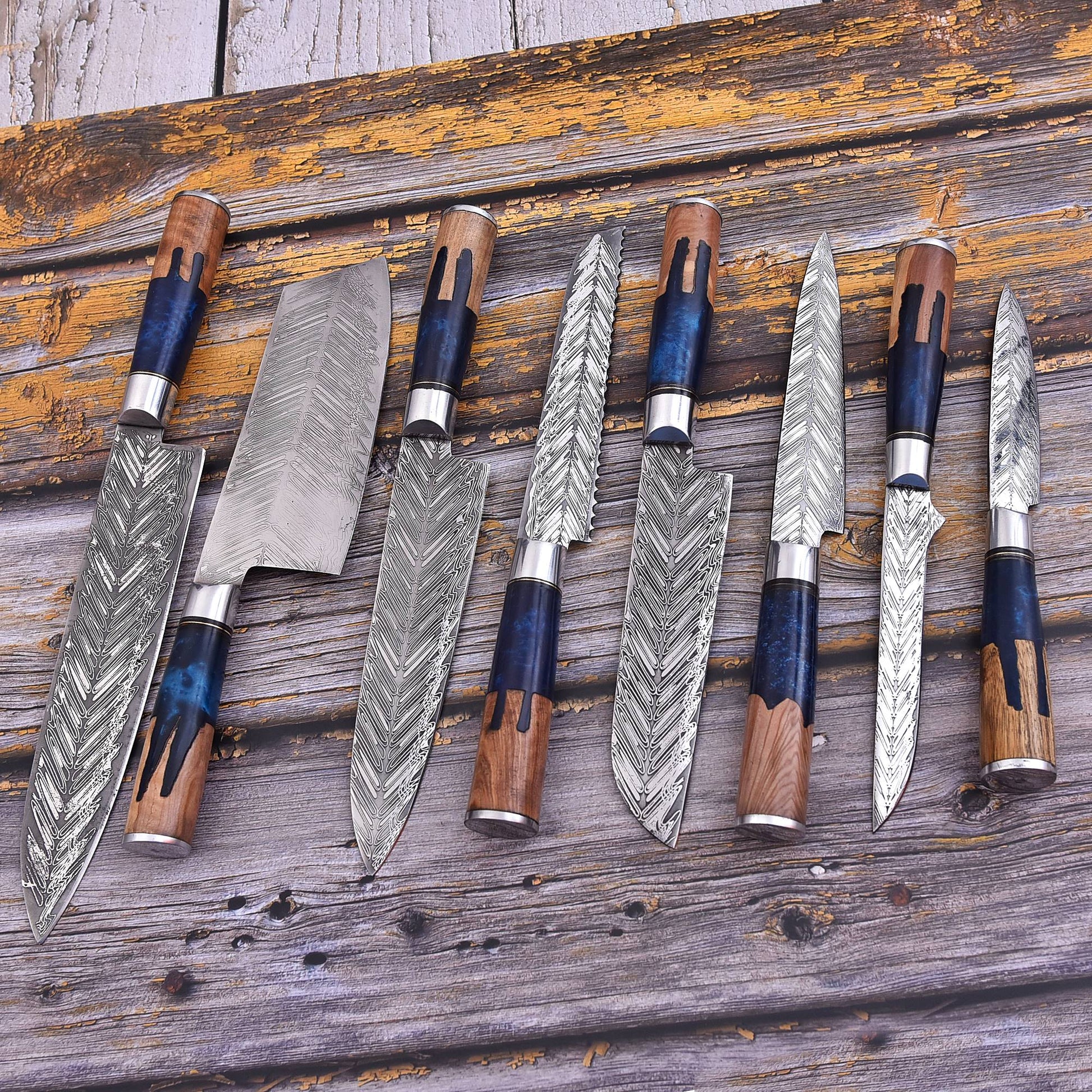Seven handmade kitchen knives with patterned Damascus steel blades and wooden handles with blue inlays, arranged on a rustic weathered wooden plank background.