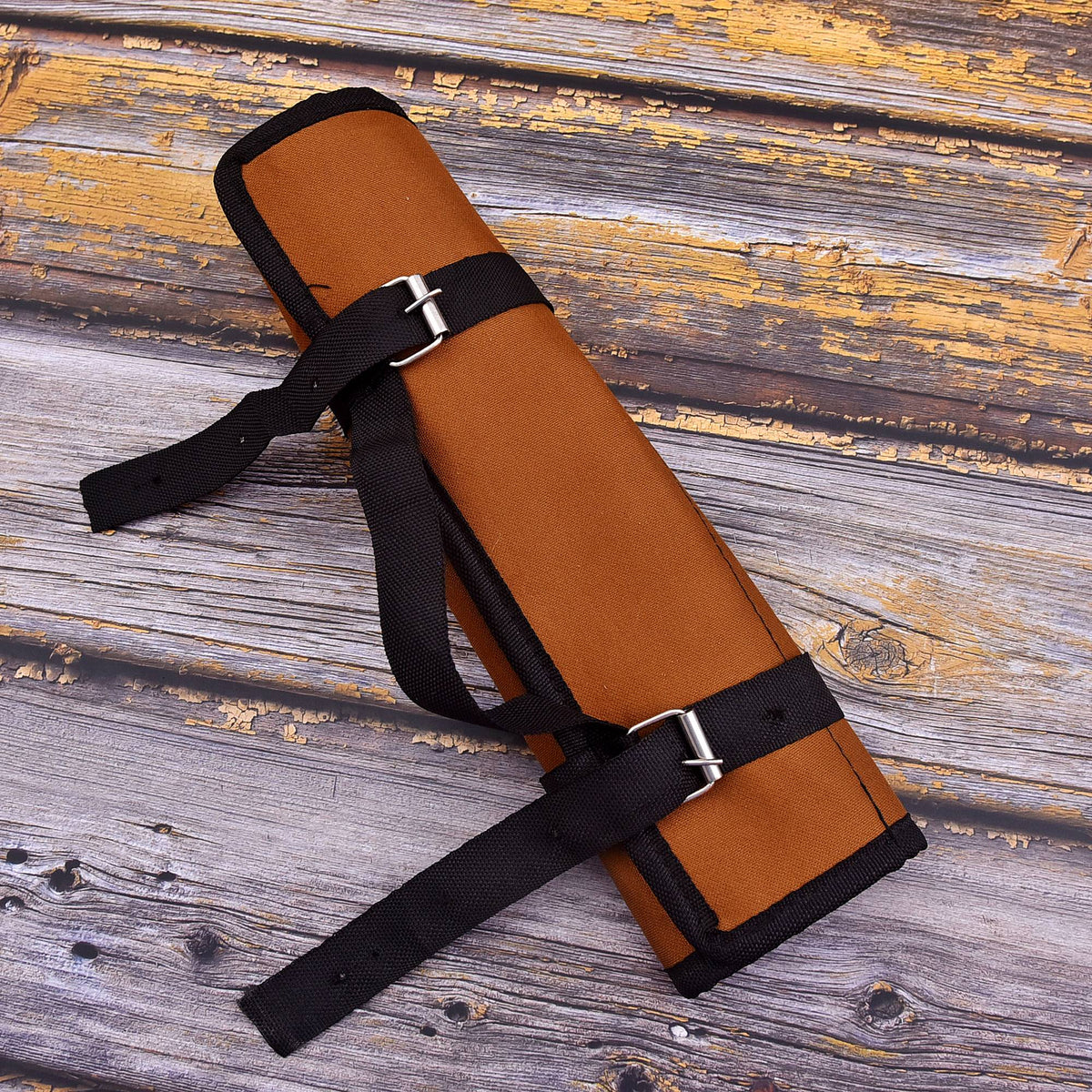 A rolled brown canvas or suede-like strap case with black webbing straps and silver buckle closures, photographed on a rustic weathered wood plank surface.