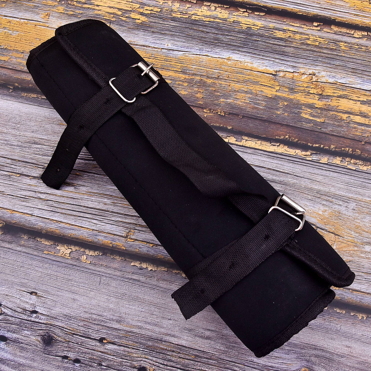 A black rolled exercise or yoga mat with adjustable black straps and silver metal buckles, placed diagonally on a weathered gray wooden plank surface.