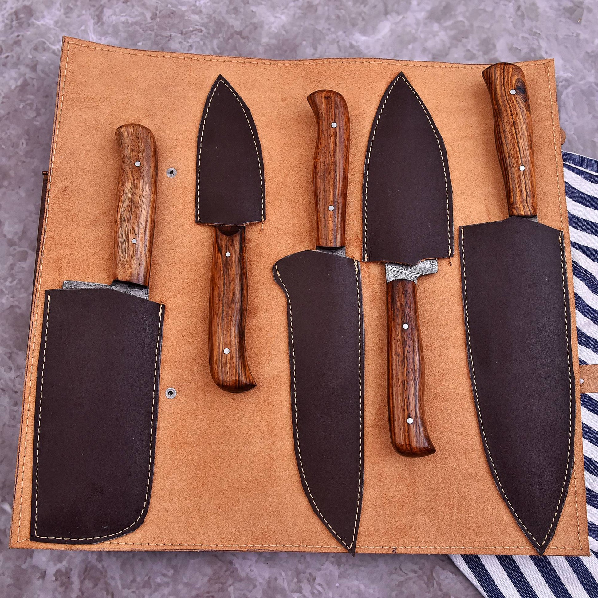 A 5-piece kitchen knife set with dark brown wooden handles and matching dark brown leather sheaths, displayed inside an open tan leather roll-up case on a gray textured surface.