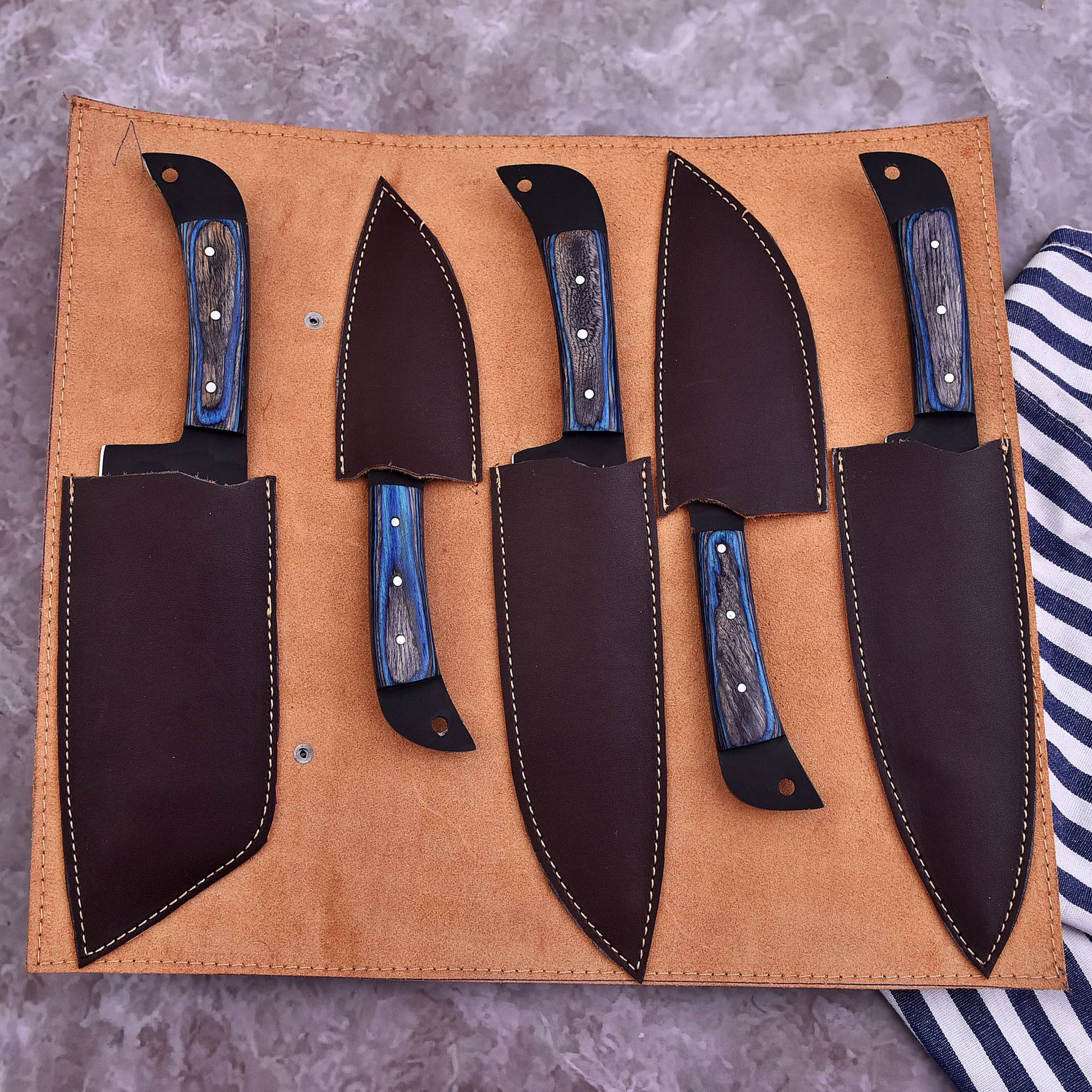 A set of five kitchen knives with blue wood handles and black blades, displayed in a tan leather roll-up sheath on a gray stone surface.
